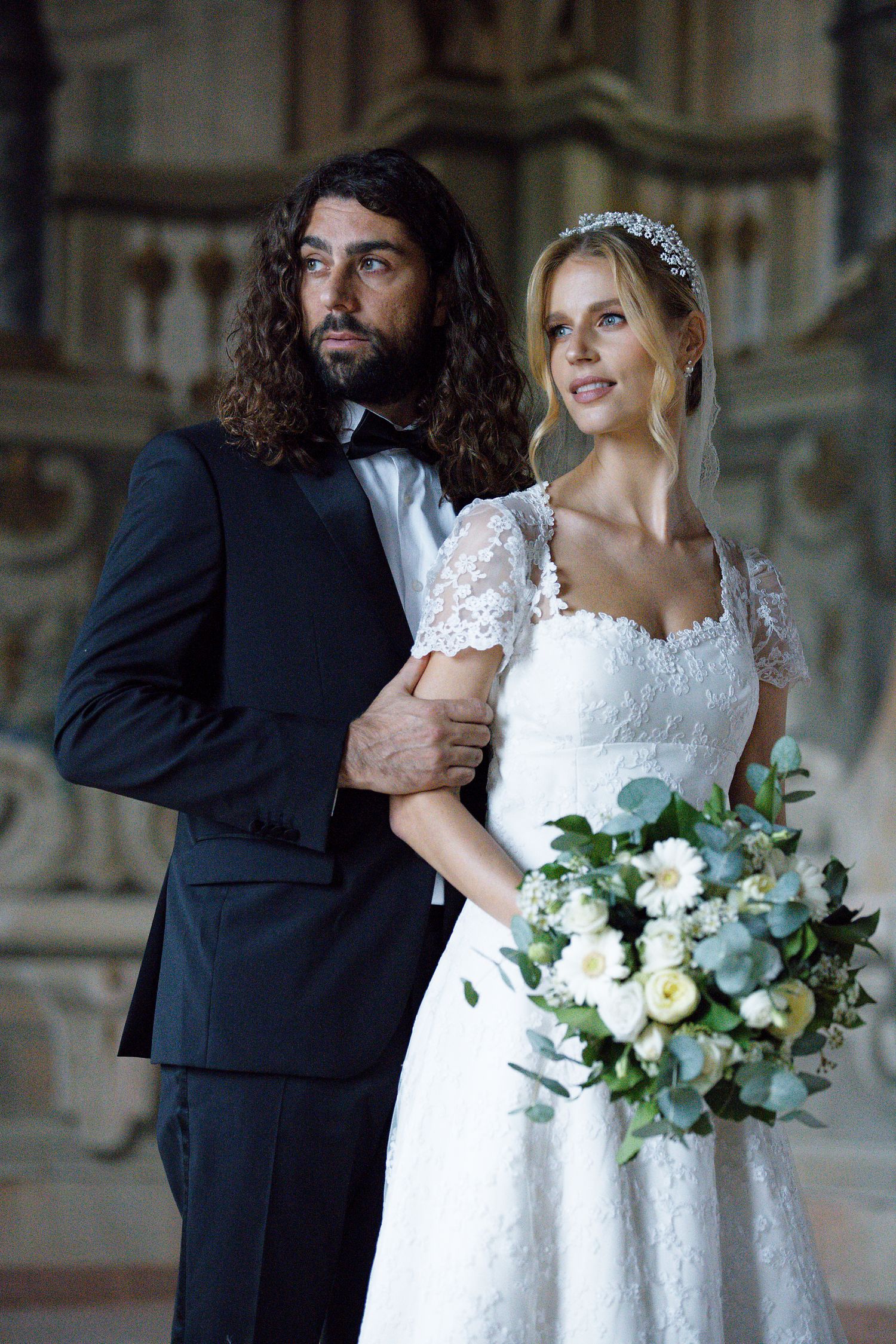 A newlywed couple poses together, the bride in a white lace dress with bouquet and the groom in a dark suit.
