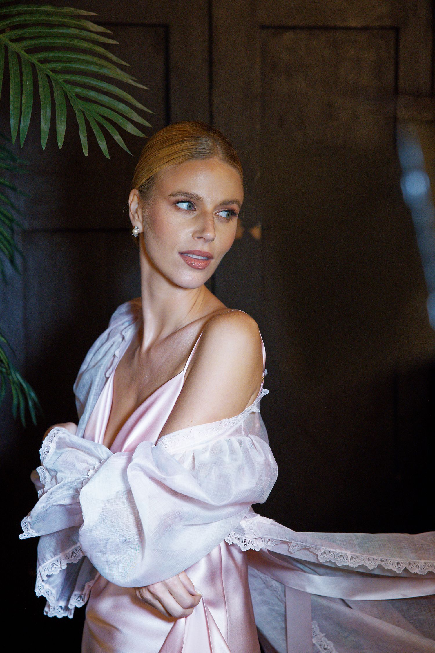 Woman in pink silk dress and white lace robe poses elegantly against dark backdrop with tropical palm fronds.