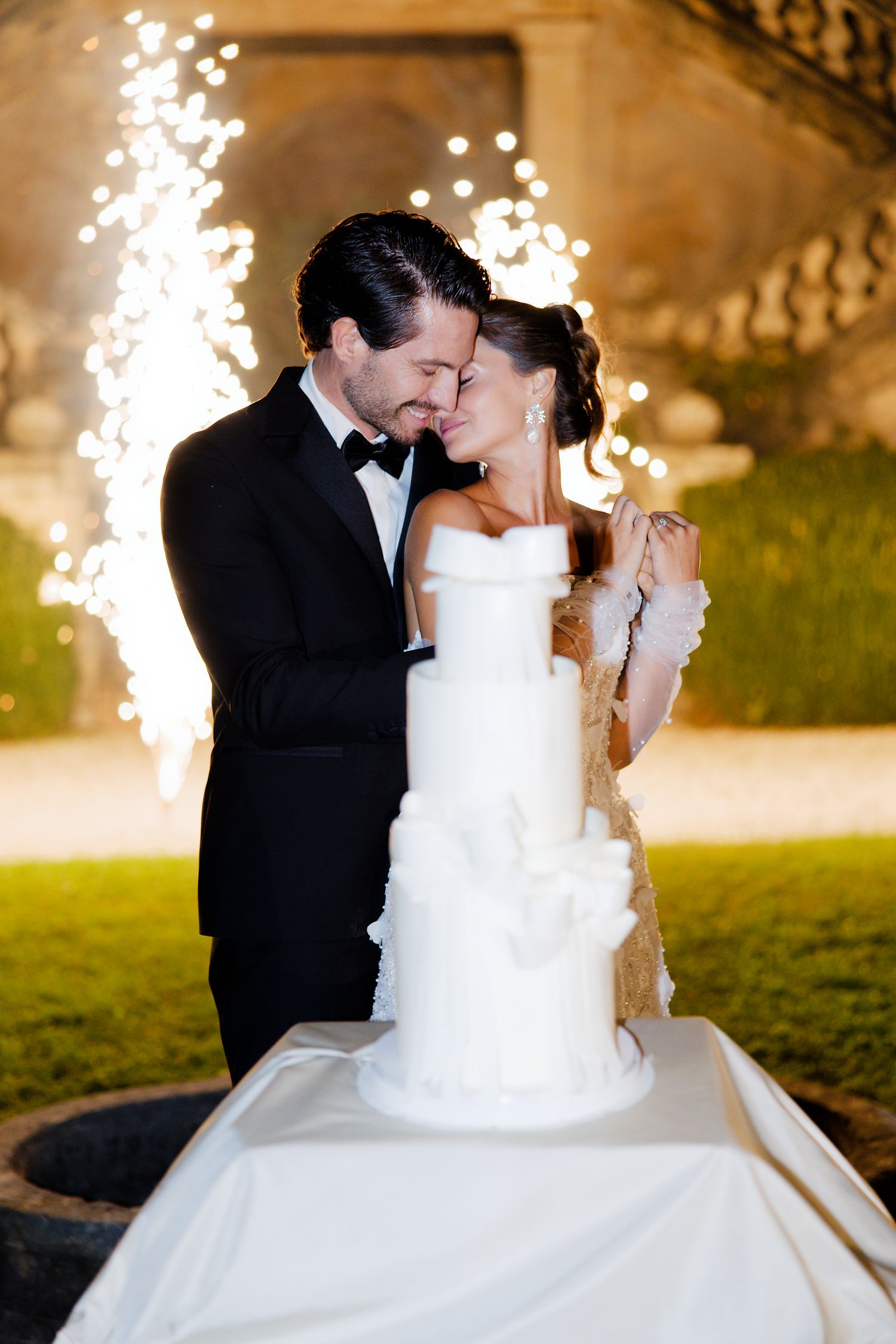 Couple cuts elegant white wedding cake with sparkler fountains illuminating the night behind them.