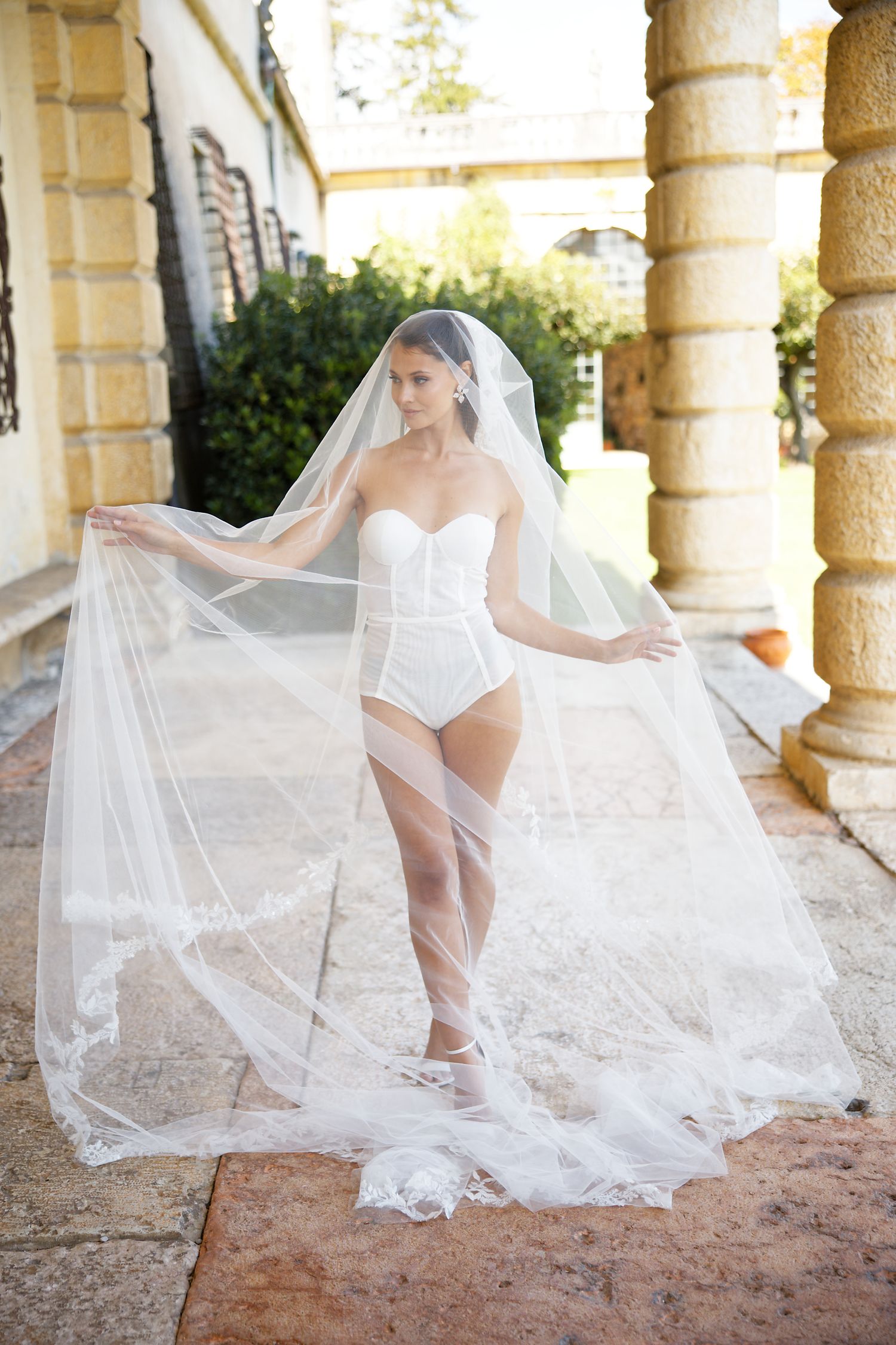 A bride in a white bodysuit poses with a flowing veil in a Mediterranean-style courtyard.