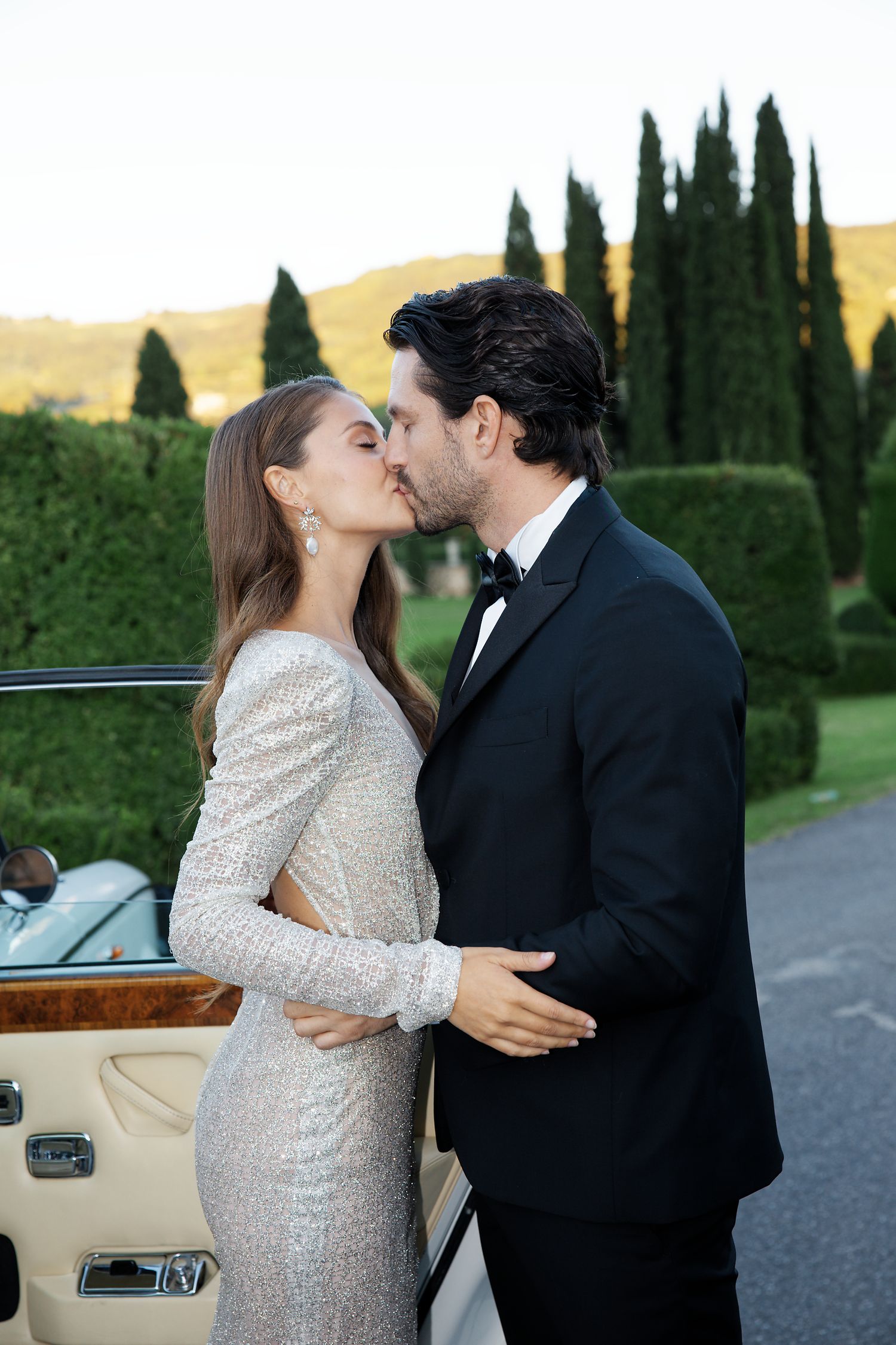 A romantic moment shared beside a vintage car with cypress trees in the Italian countryside.