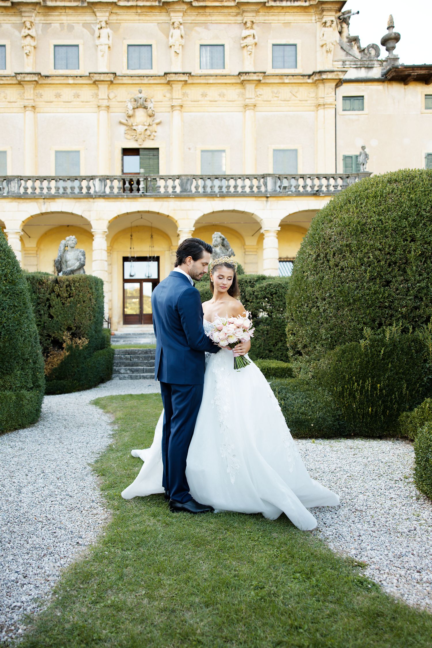 A wedding scene on a gravel path surrounded by manicured hedges at a grand Italian-style villa.