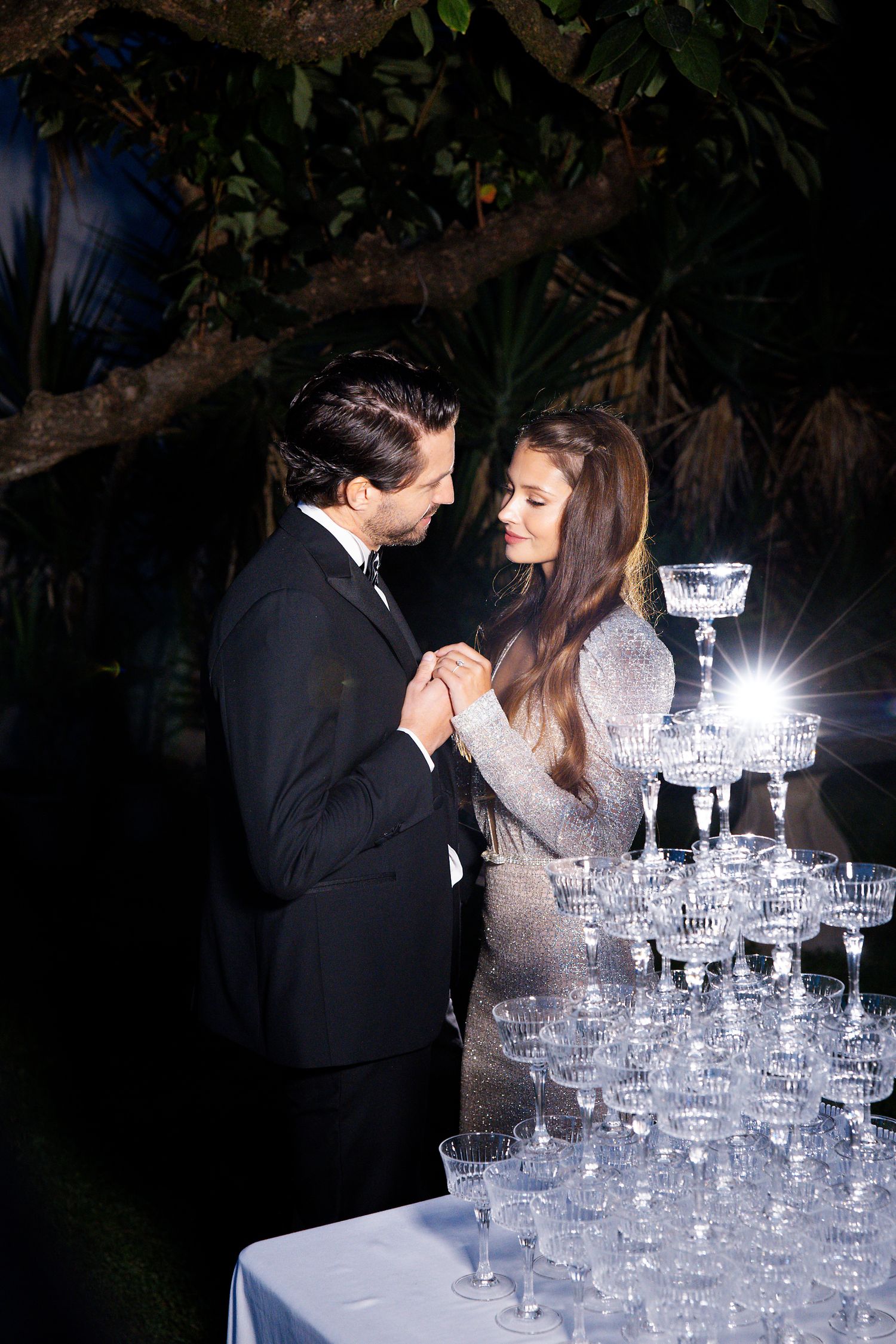 Couple stands by tiered champagne tower at evening wedding reception.