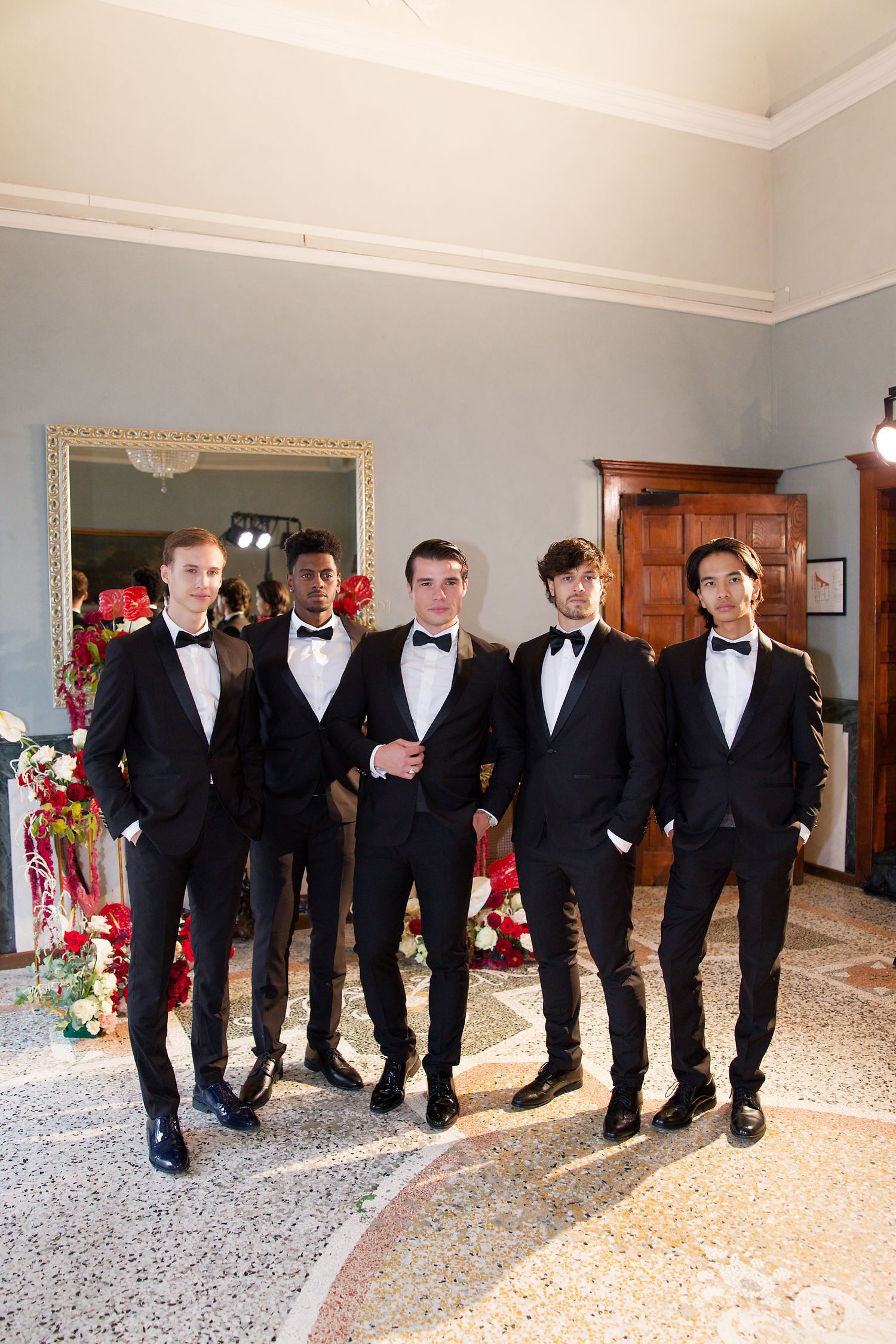 Groomsmen in black tuxedos and bowties stand together in an elegant room with ornate decor.