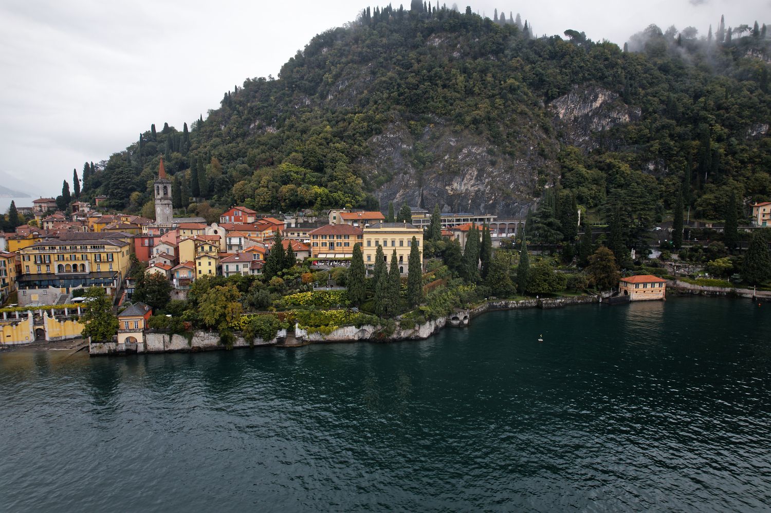 A scenic lakeside village with colorful buildings nestled along Lake Como in Italy beneath a forested mountain.