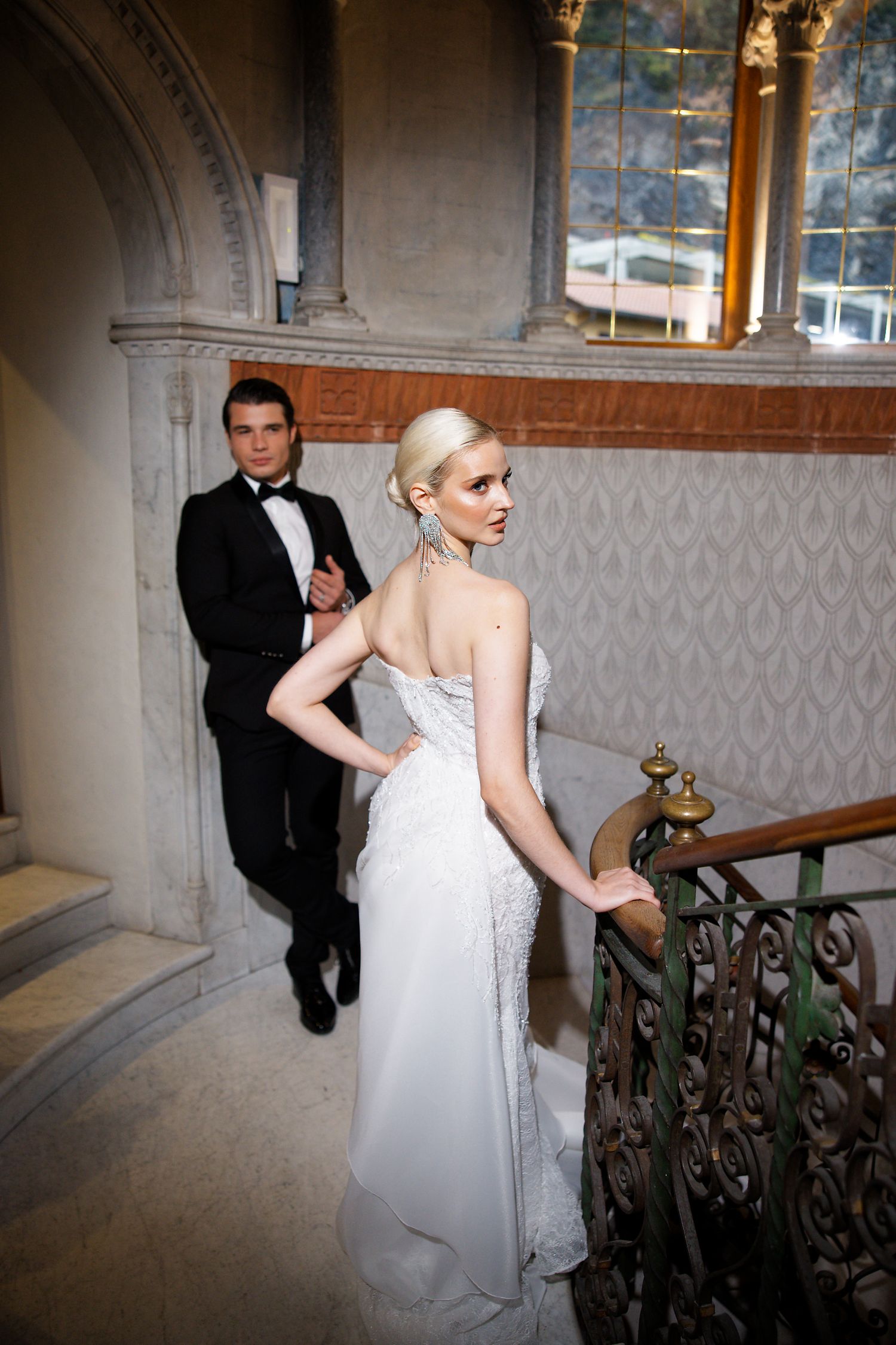 A couple in formal attire pose on an elegant staircase with architectural details.