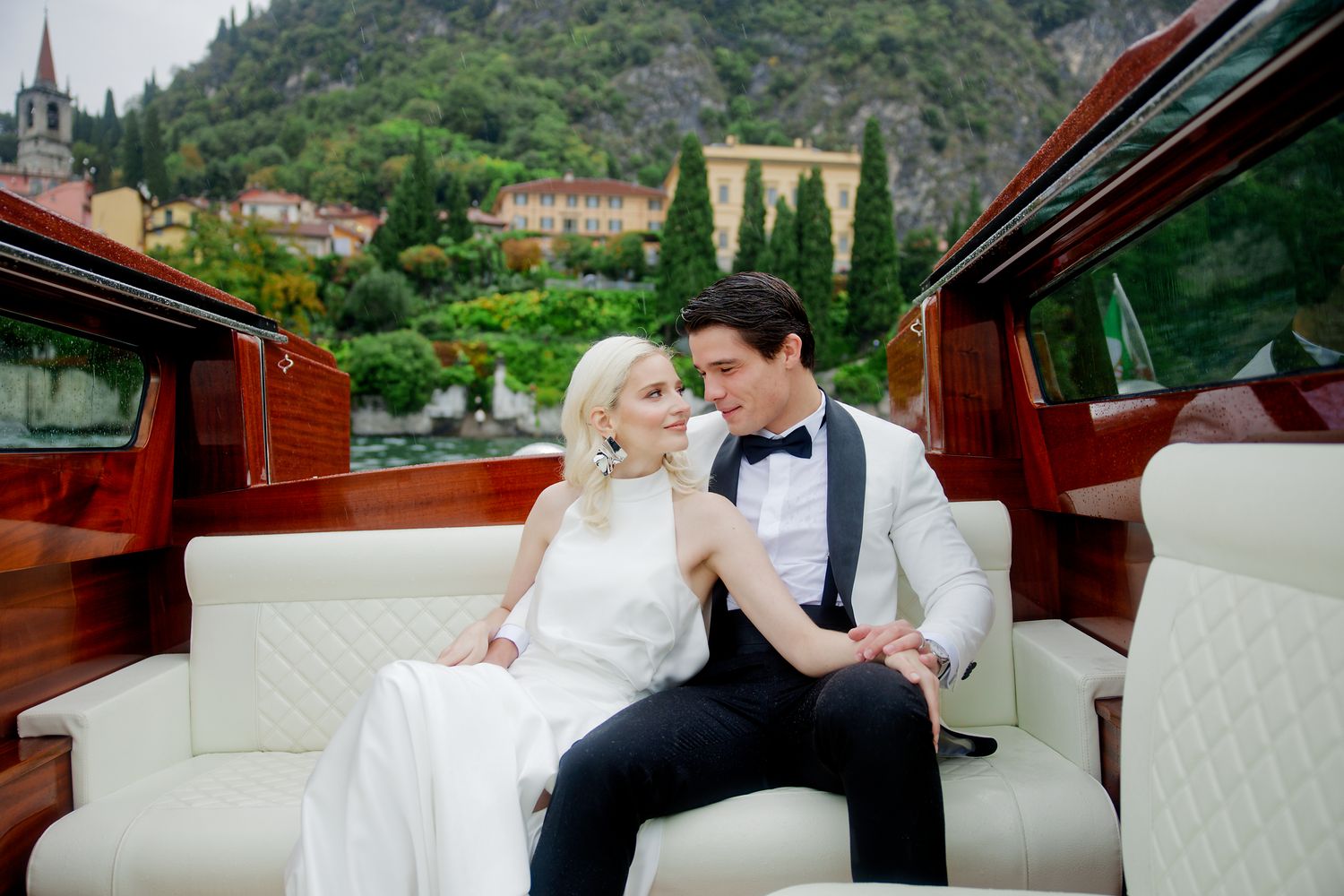 A couple in wedding attire share an intimate moment while seated in a vintage boat with scenic hills in the background.