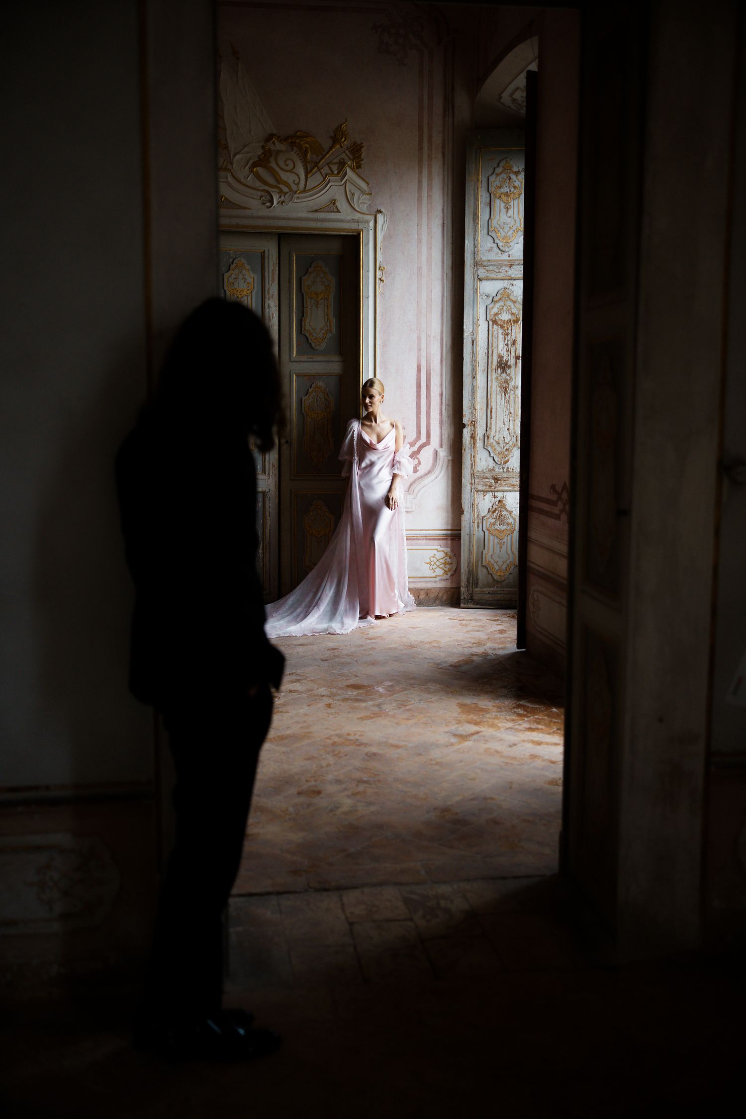 Dramatic boudoir photo showing silhouette observing figure in pink dress through ornate vintage doorway.