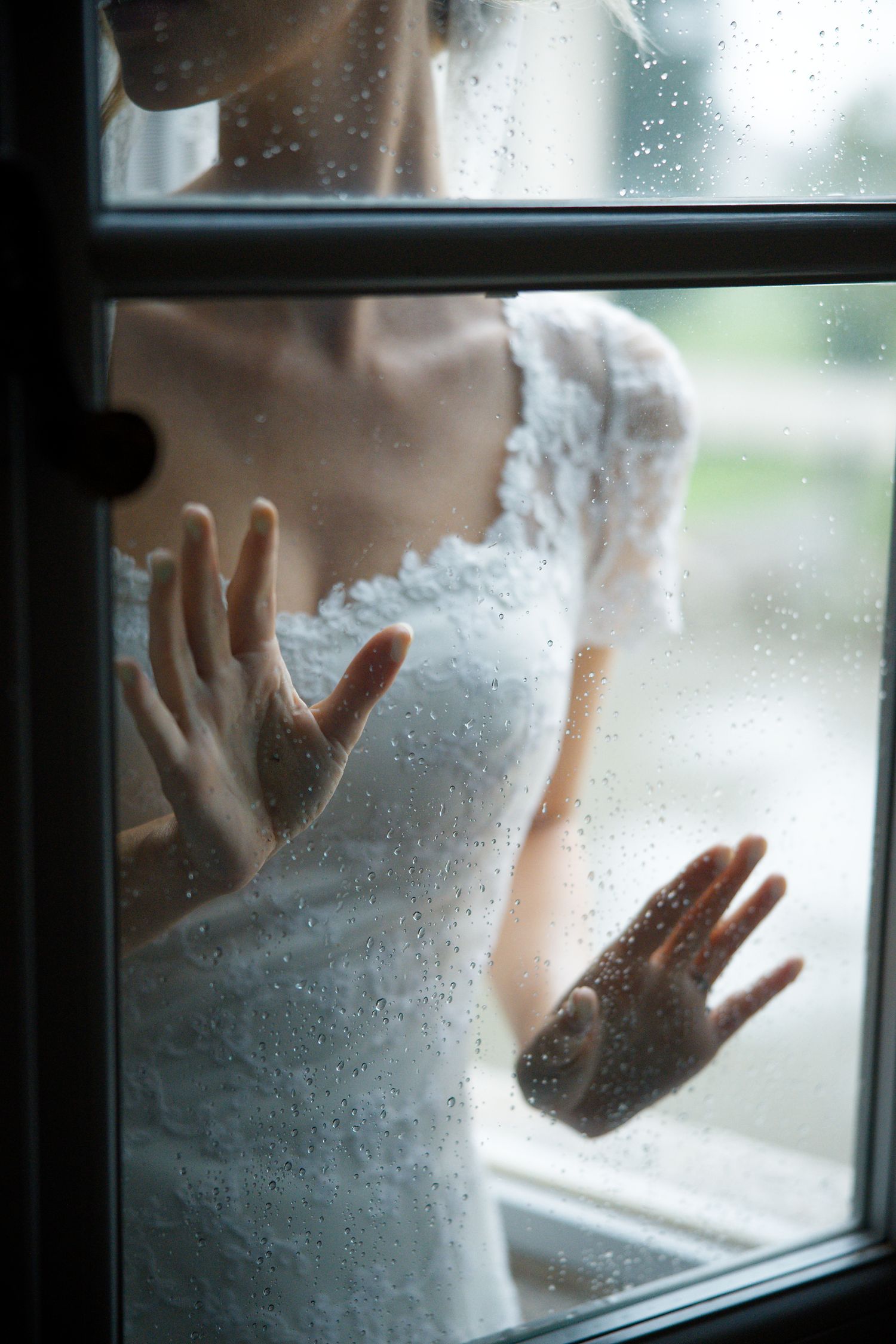 Hands press against a rain-streaked window, creating a dreamy artistic wedding photograph.