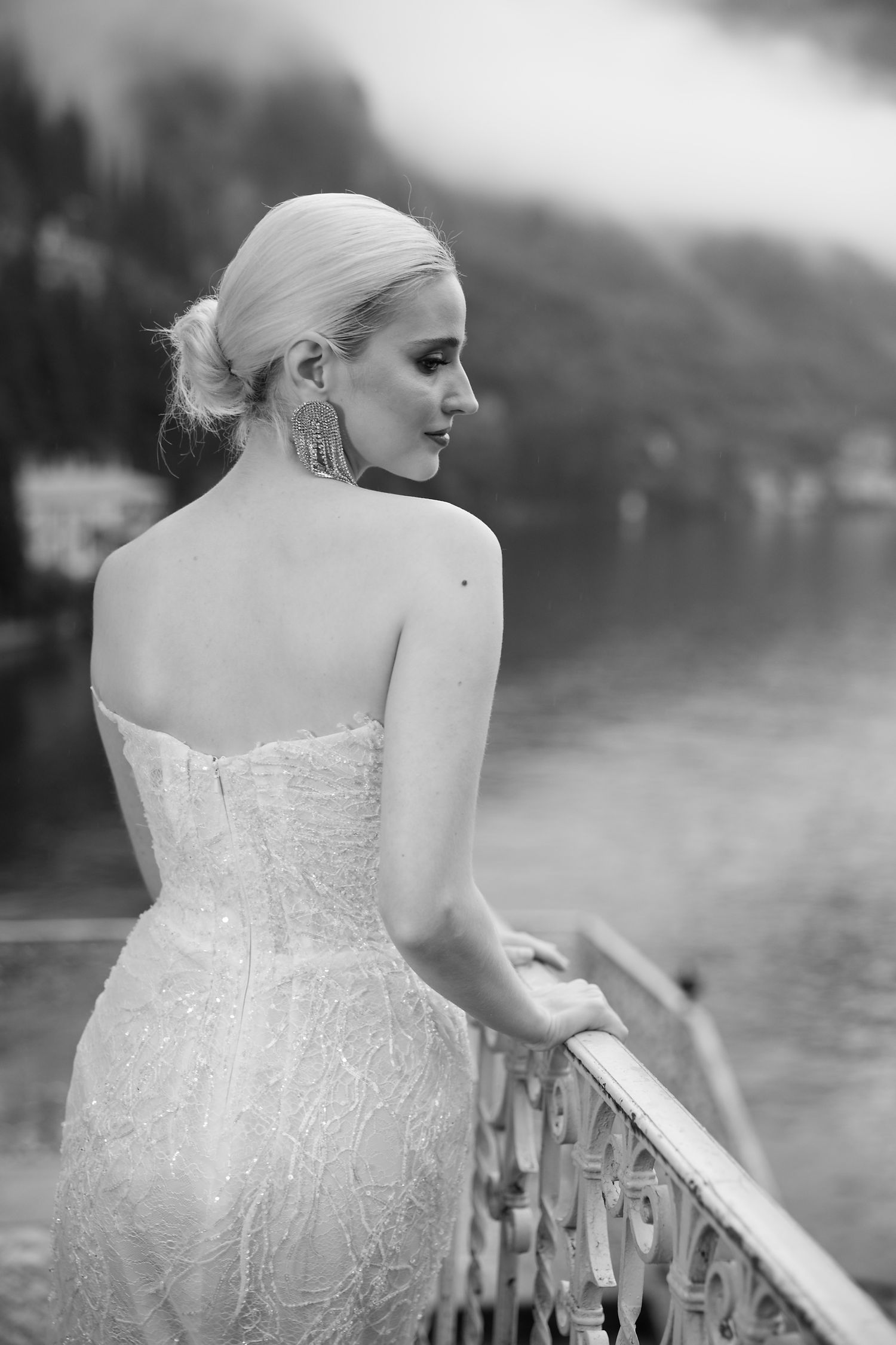 Bride in lace gown looks over shoulder while standing on lakeside balcony in black and white portrait.