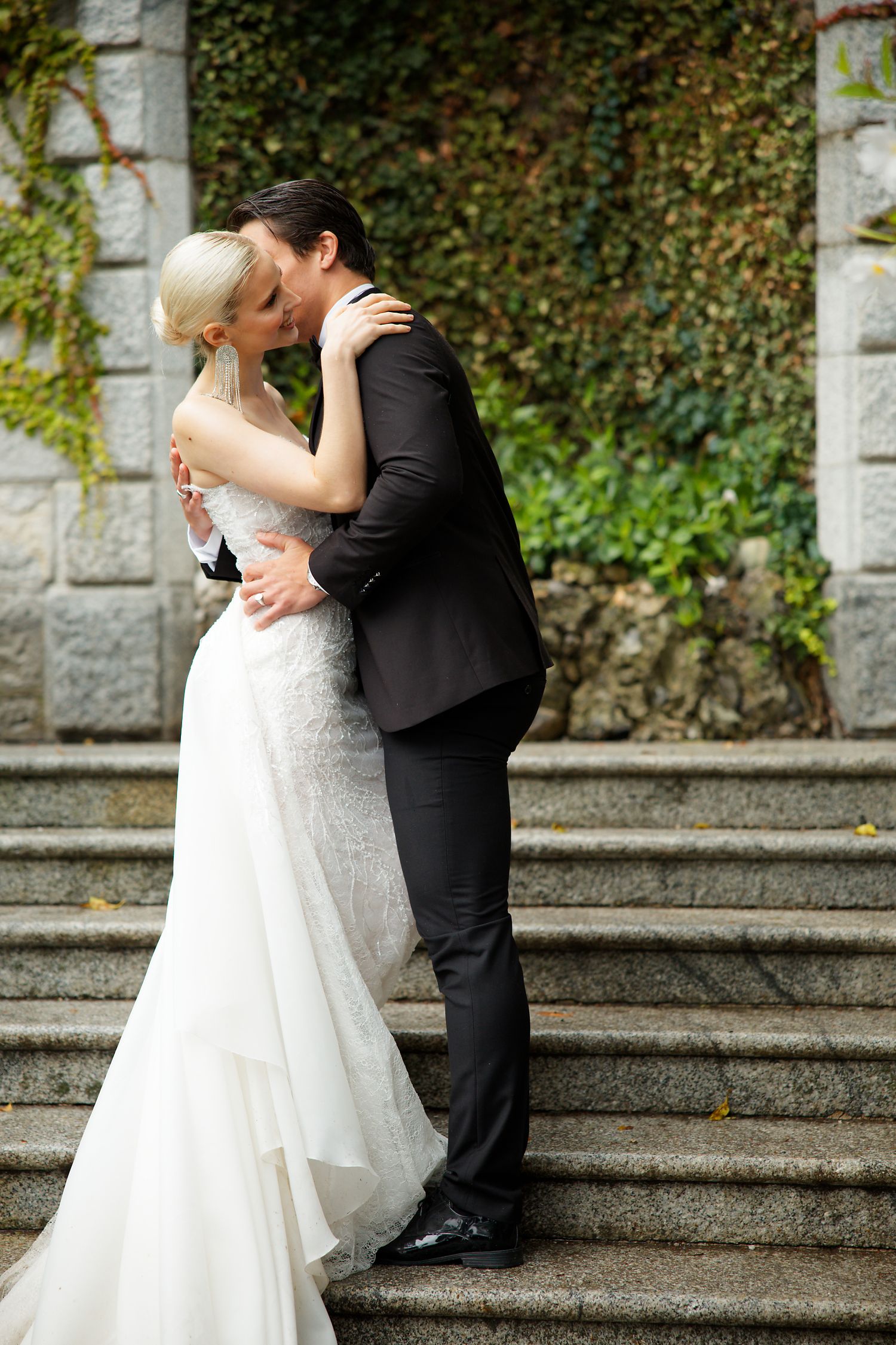 A romantic wedding couple sharing a kiss on stone steps surrounded by ivy-covered walls.