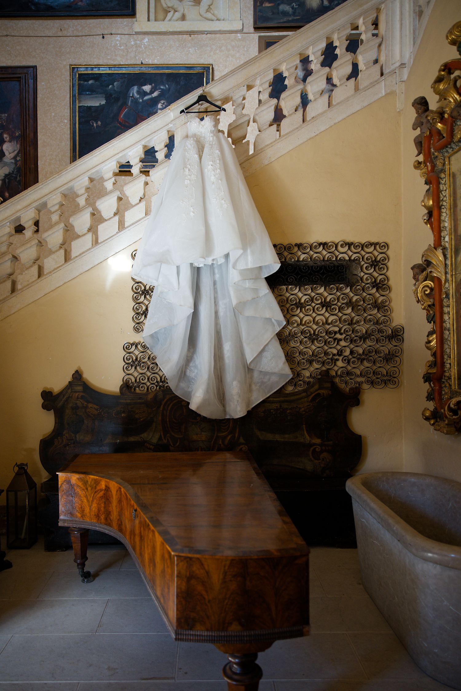 Classic white wedding dress displayed on hanger against ornate staircase in vintage interior.