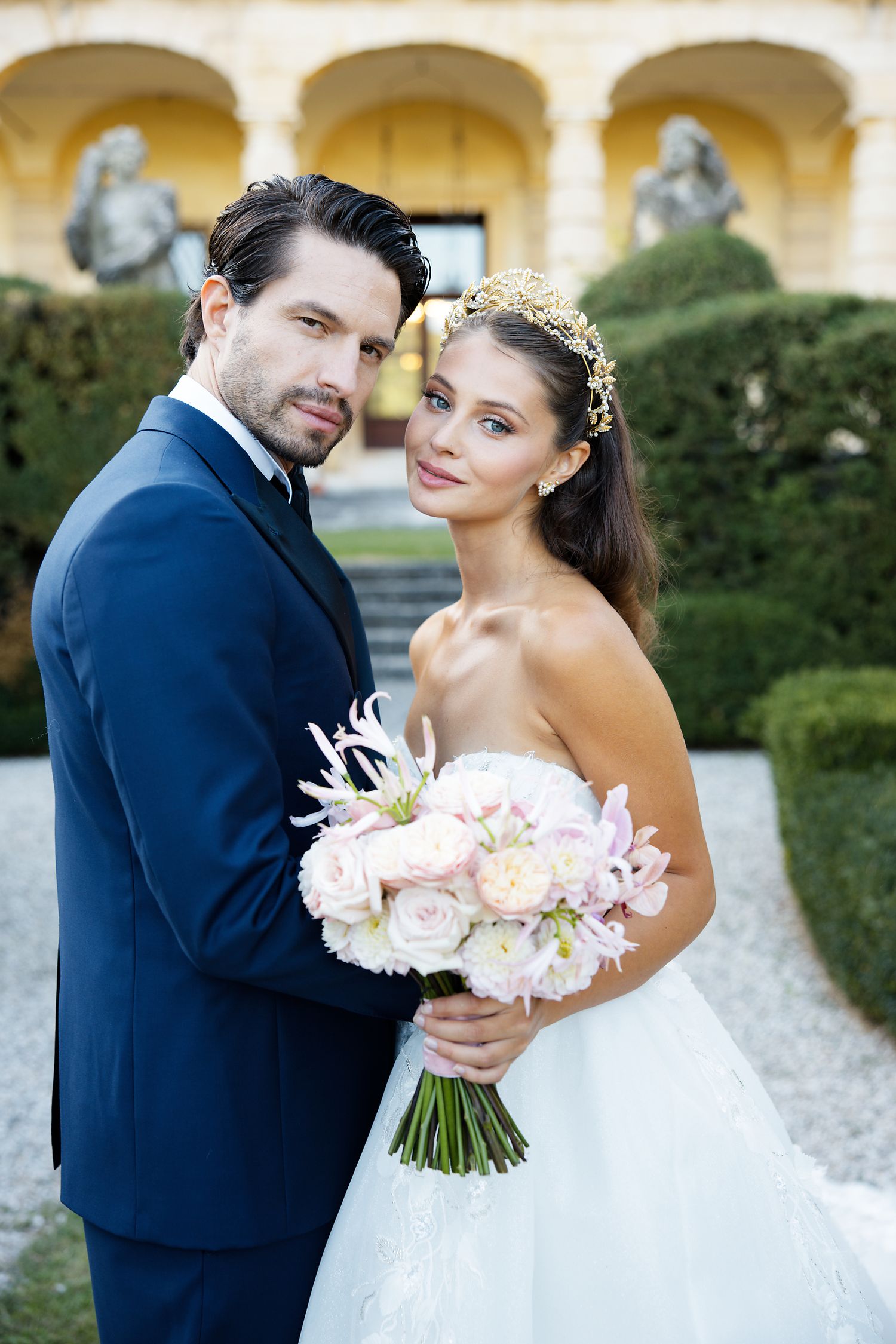 Couple in formal wedding attire poses in garden setting with pink bouquet.