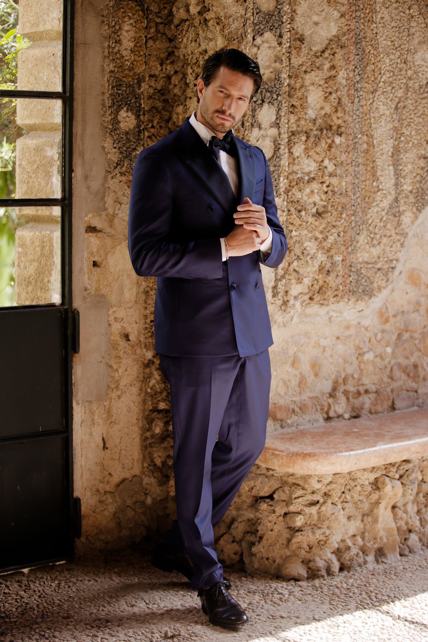 Groom in navy suit adjusts cuffs while standing against textured stone wall.