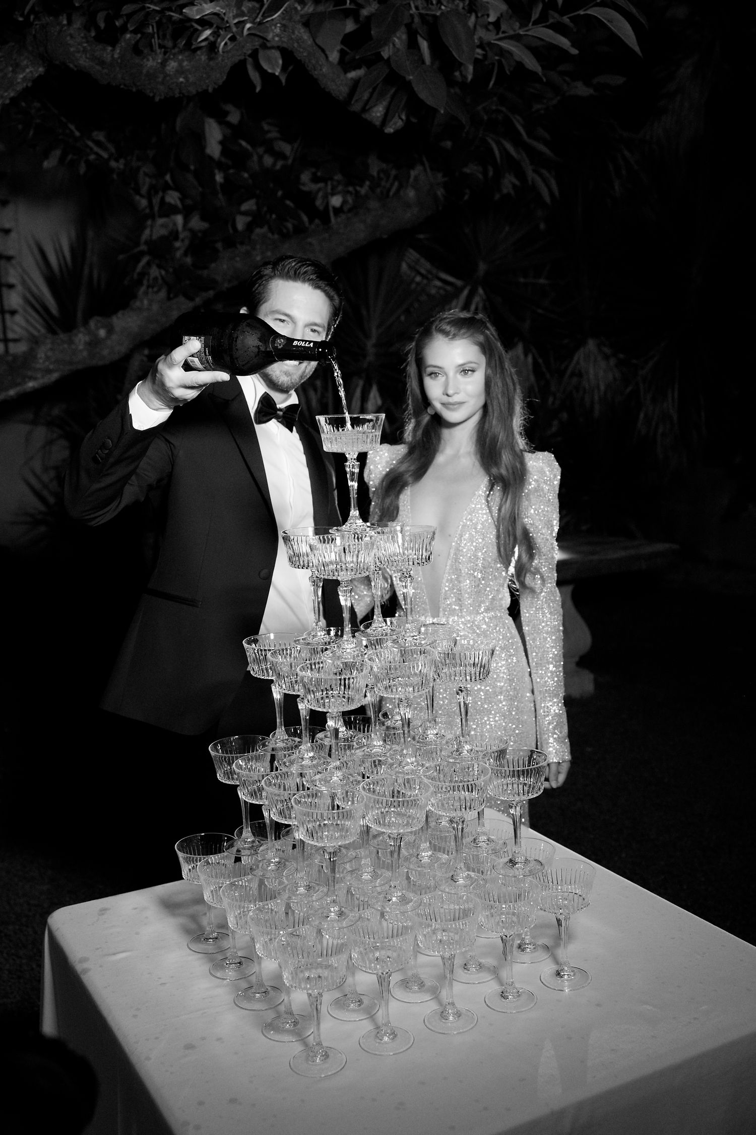 Wedding couple pouring champagne into tower of crystal glasses at outdoor reception.