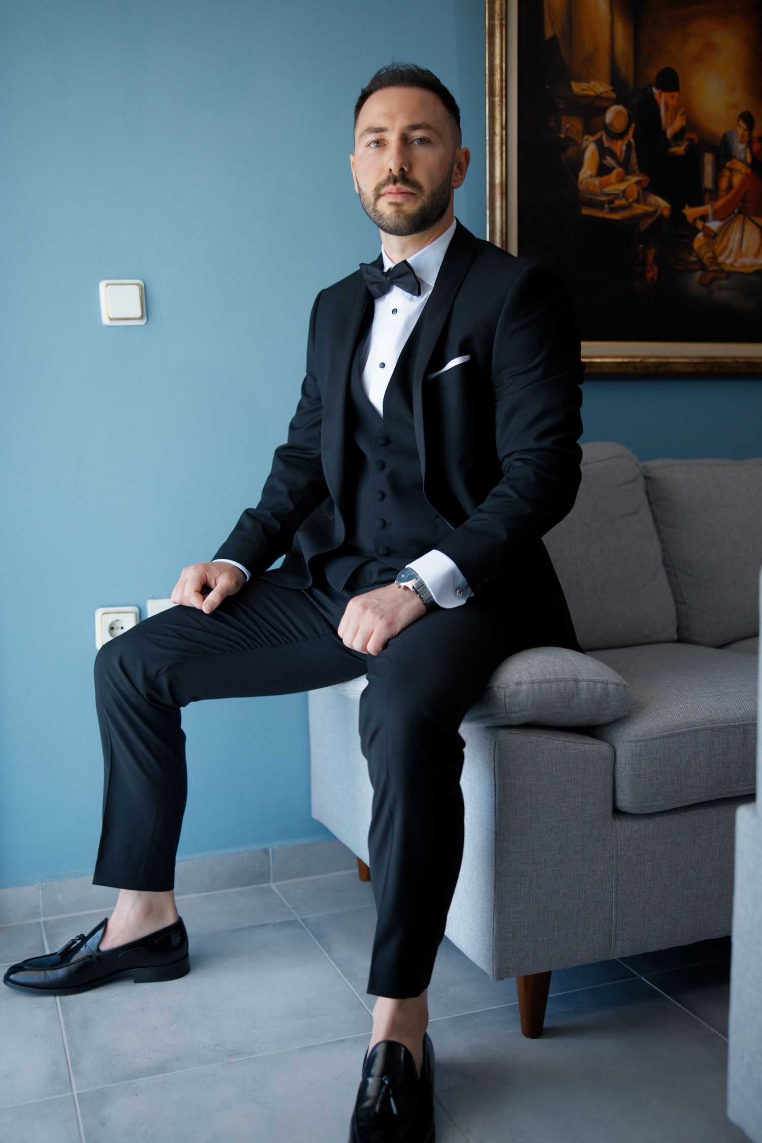 Elegant groom in black tuxedo and bow tie sits on grey sofa against blue wall with classical painting.