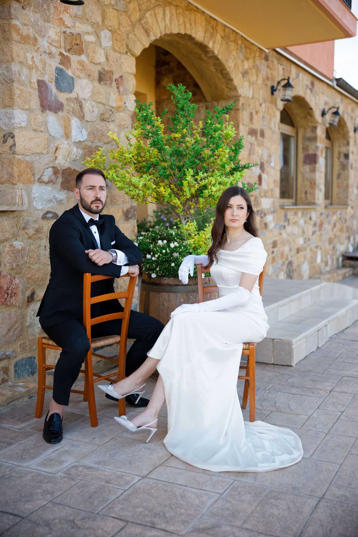A bride and groom seated on wooden chairs outside a rustic stone building during their wedding.