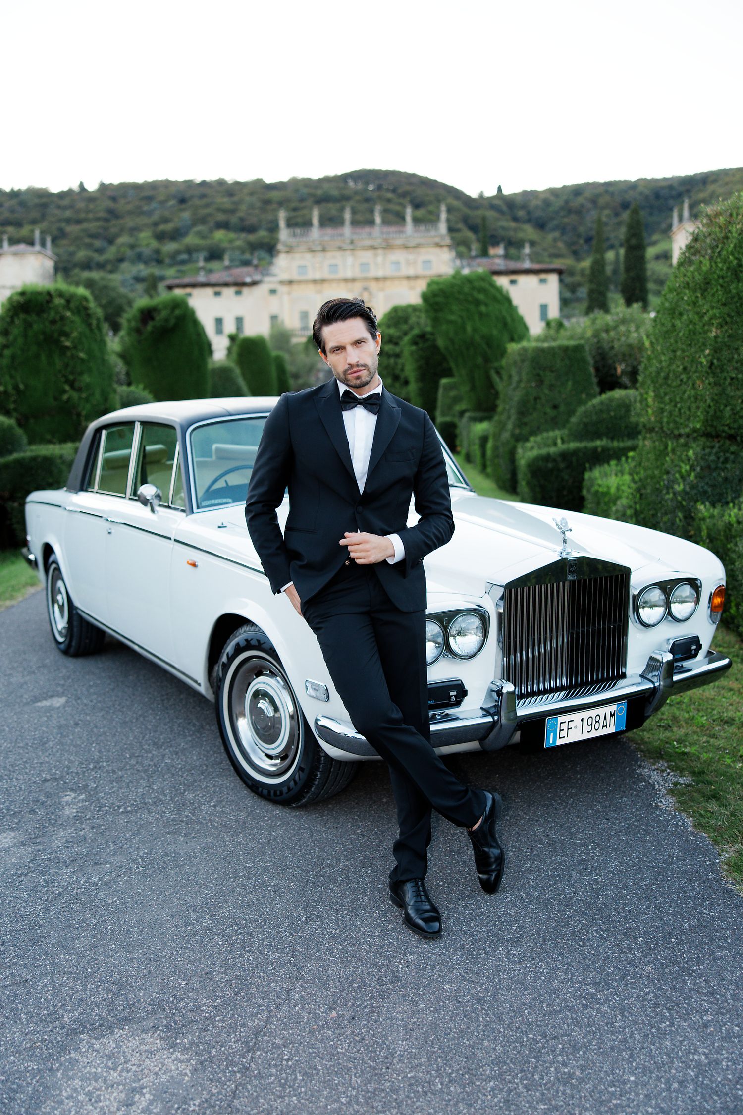Elegant classic white Rolls-Royce car parked in front of a grand Italian villa with manicured topiary gardens.