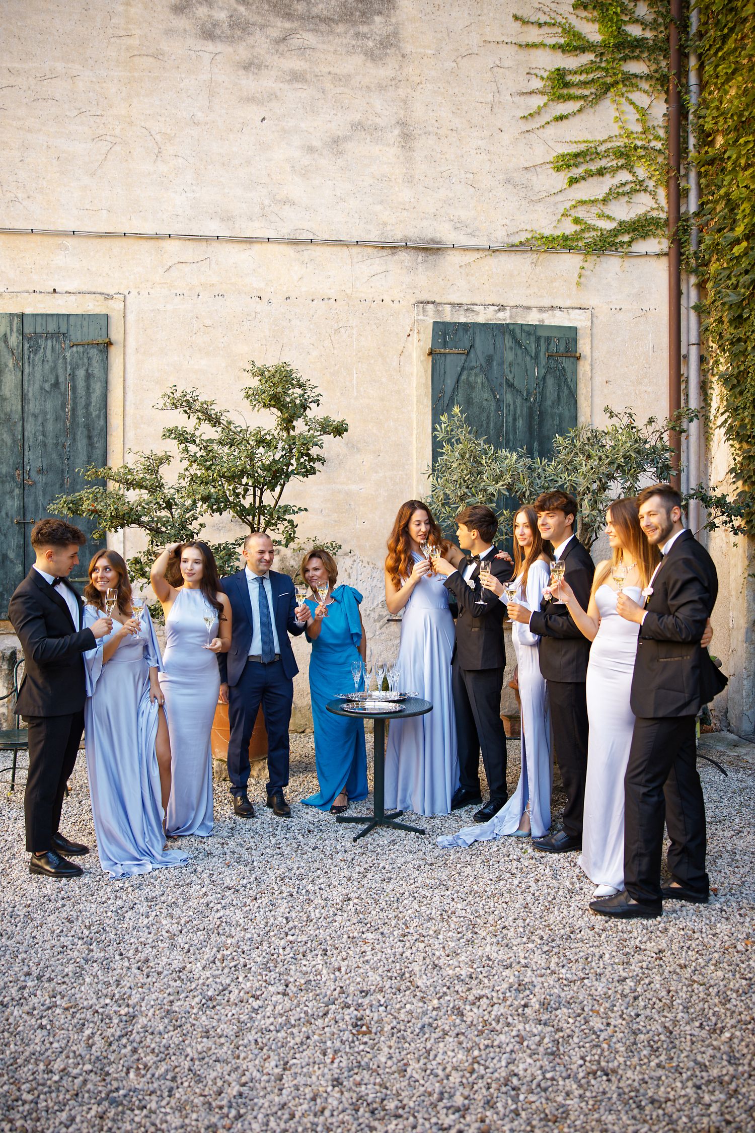 Wedding party in light blue dresses gathers on gravel outside rustic building with blue doors.