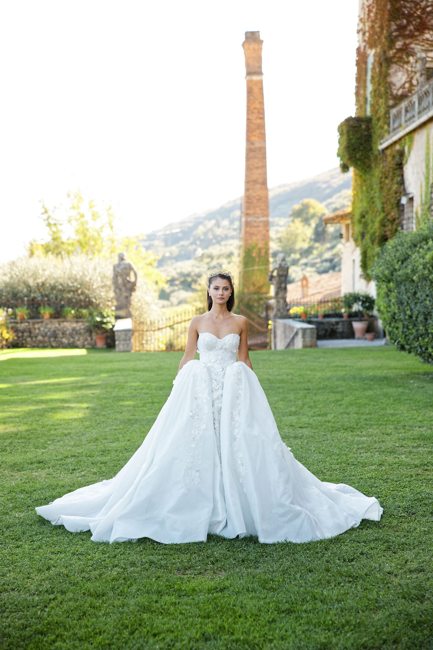 Stunning white wedding gown with flowing train photographed on manicured lawn with historic building backdrop.