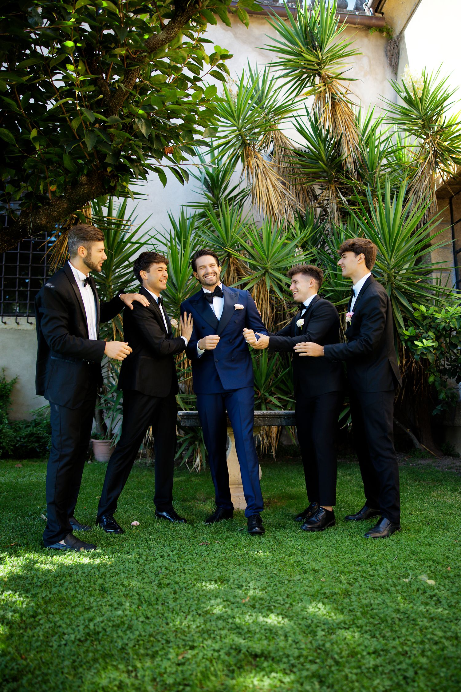 Groomsmen in tuxedos share a laughing moment in a garden setting.
