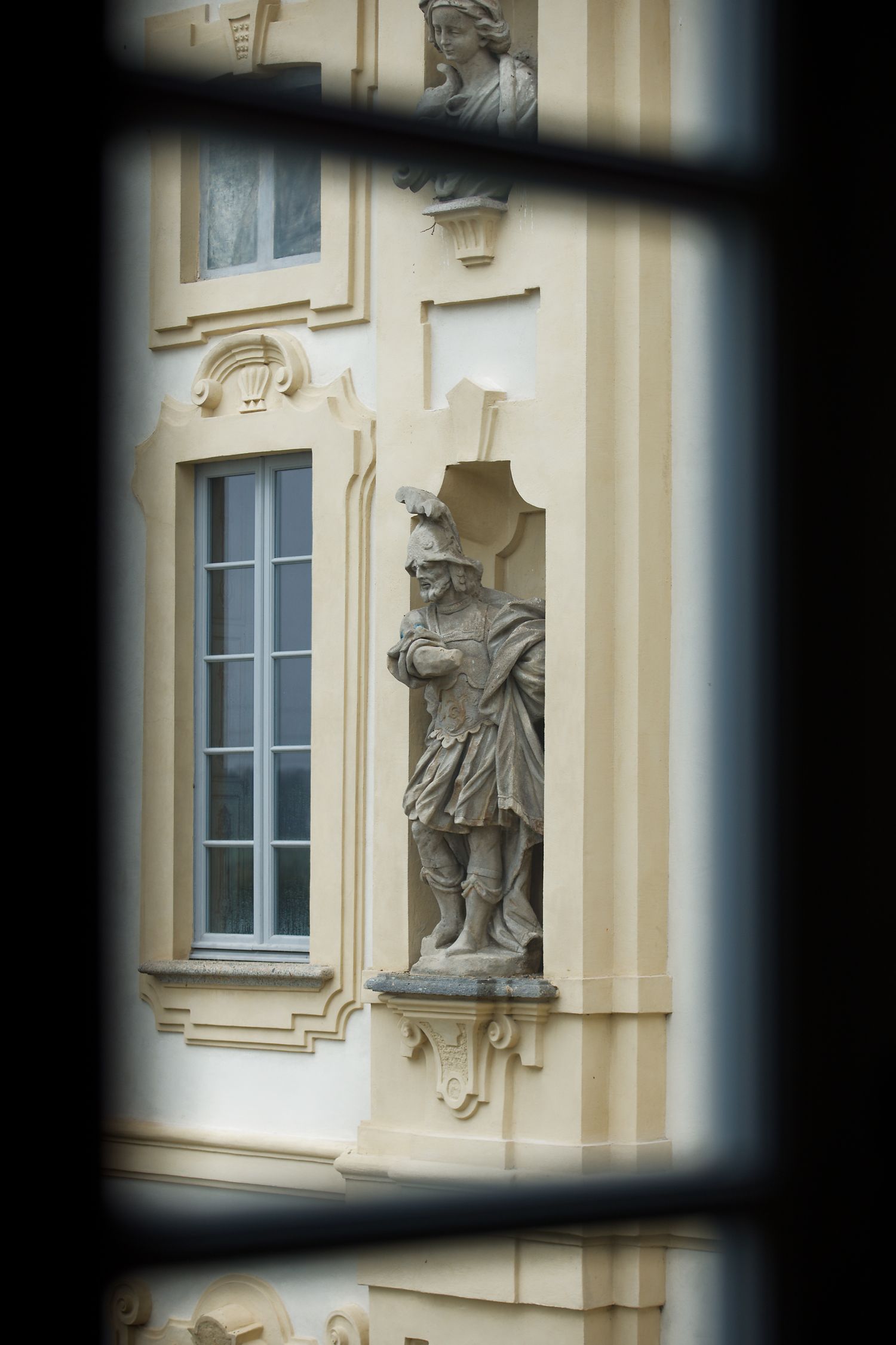 Classical stone statue viewed through window frame on beige building facade.