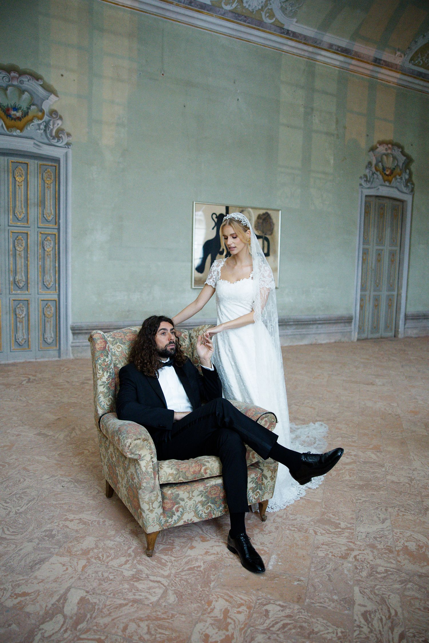 Newlyweds share a playful moment with the groom lounging in a vintage chair while the bride stands beside him.