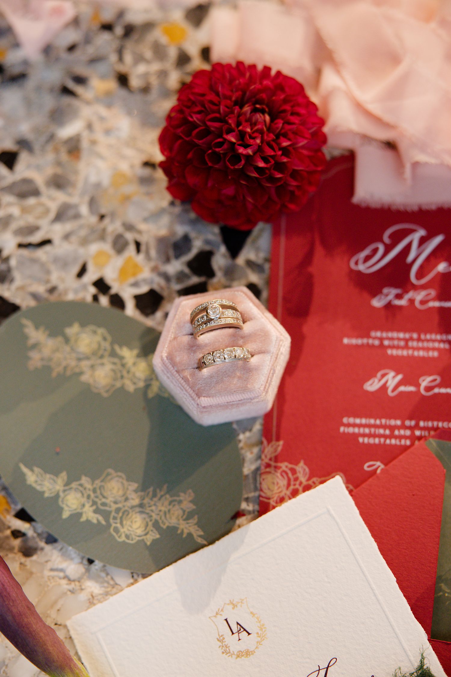 Wedding rings displayed in pink hexagonal box with red dahlia flower and invitation details.