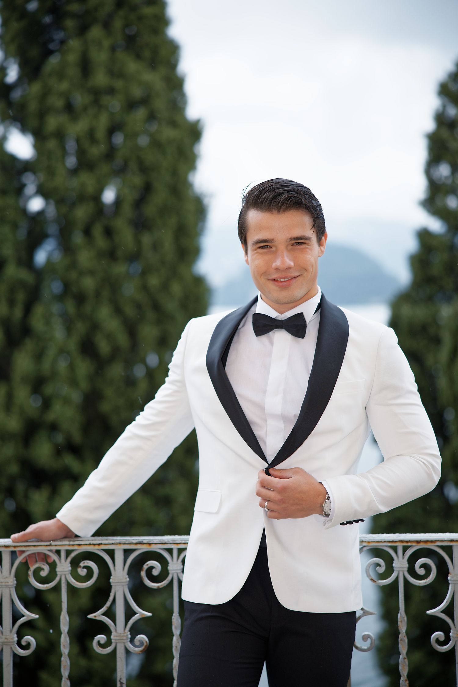 Groom in classic white tuxedo jacket and bowtie poses on ornate balcony.