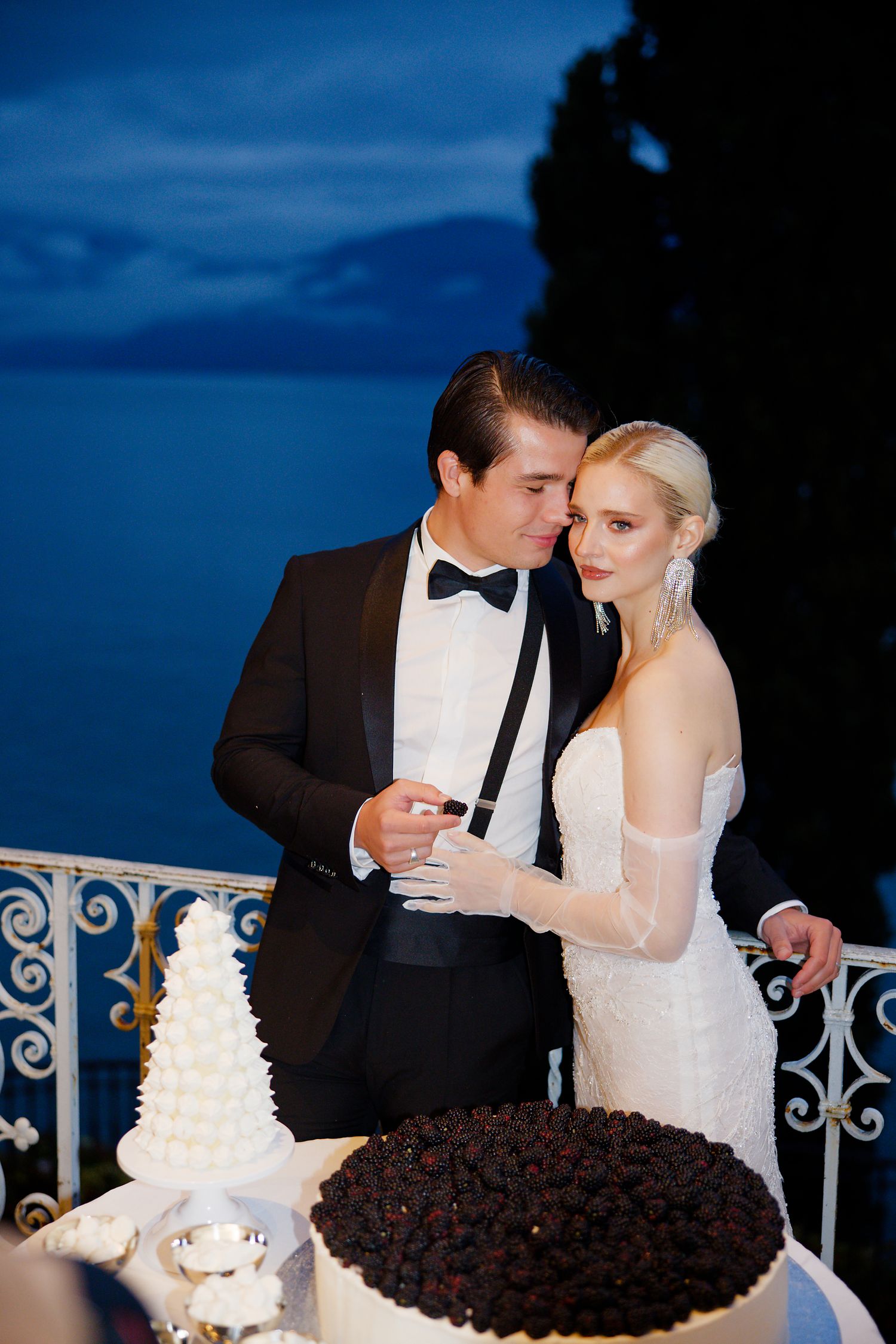 Newlyweds share an intimate moment at twilight next to an elegant wedding cake display with ornate railing.