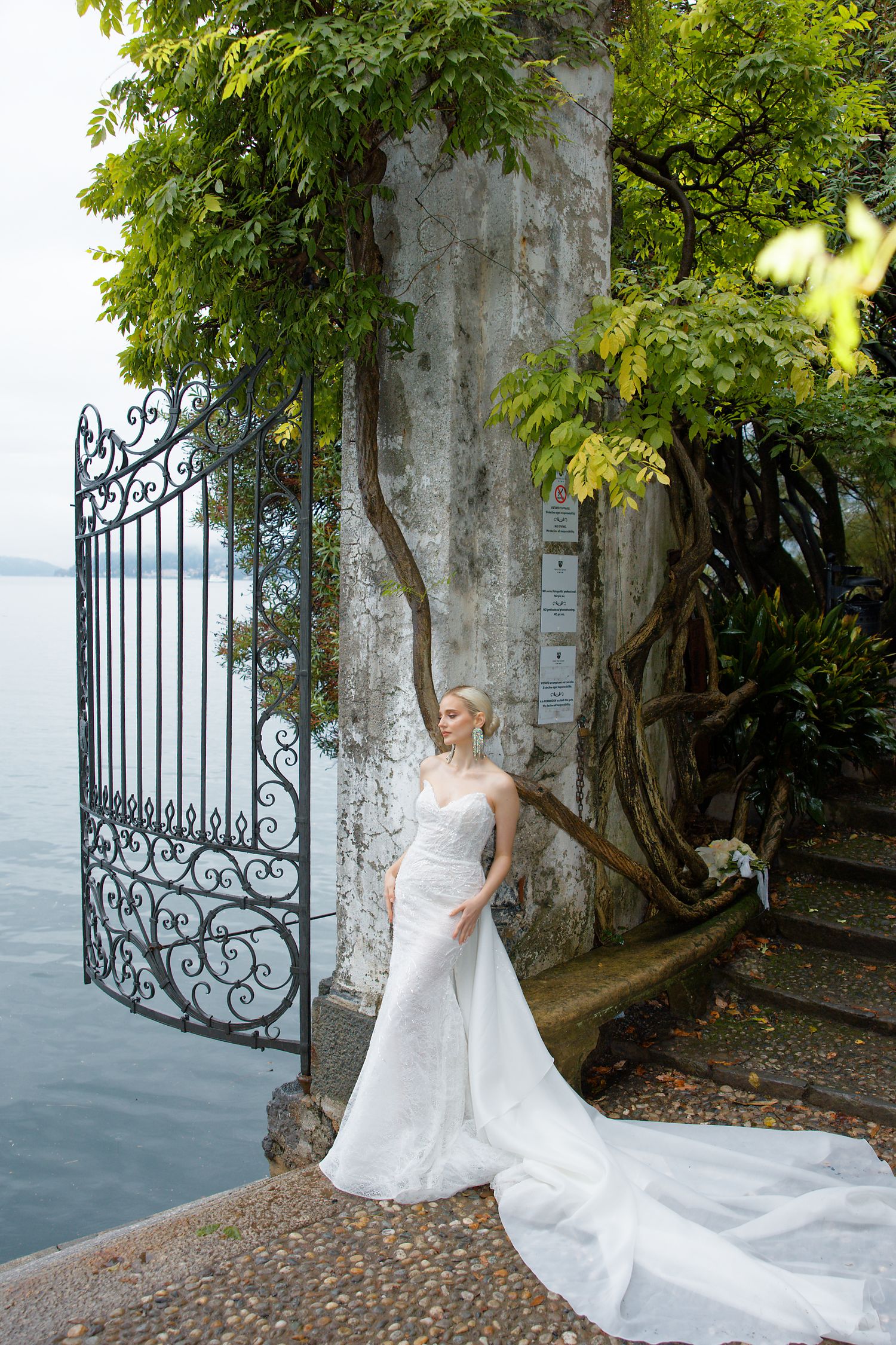 A person in a white wedding gown poses by an ornate iron gate overlooking a lake.