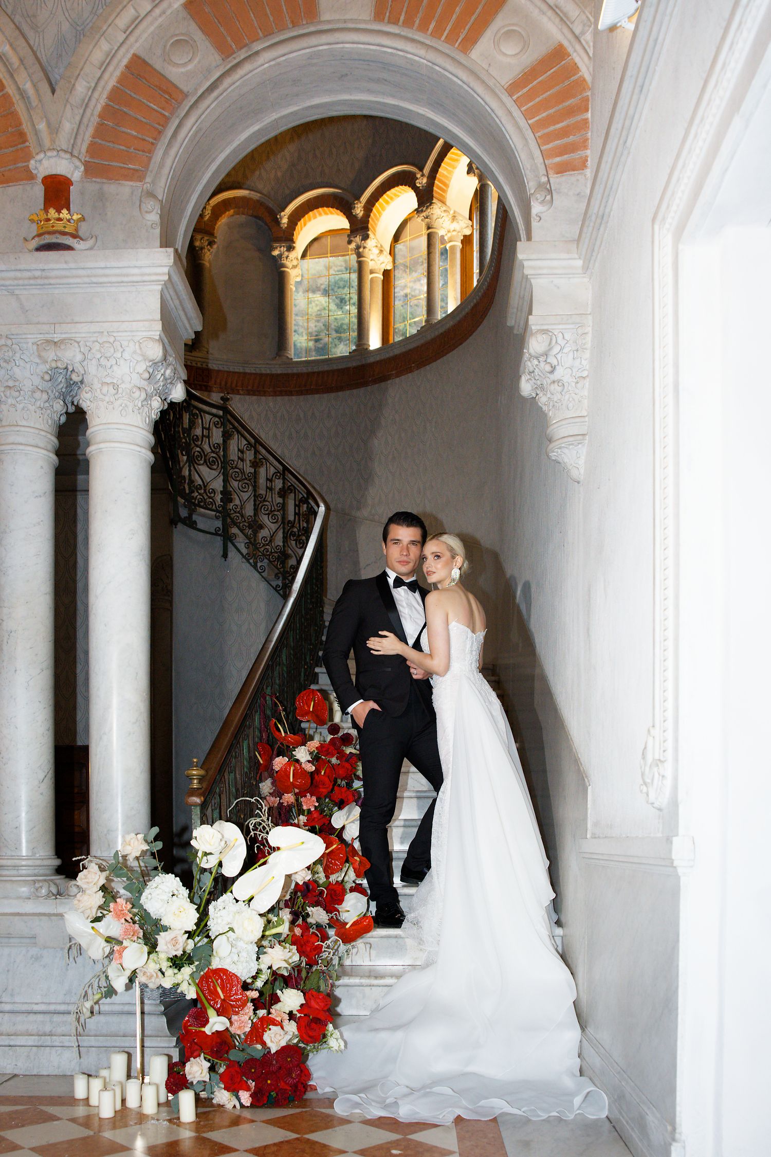 A couple stands on an ornate staircase adorned with red and white flowers under arched architecture.