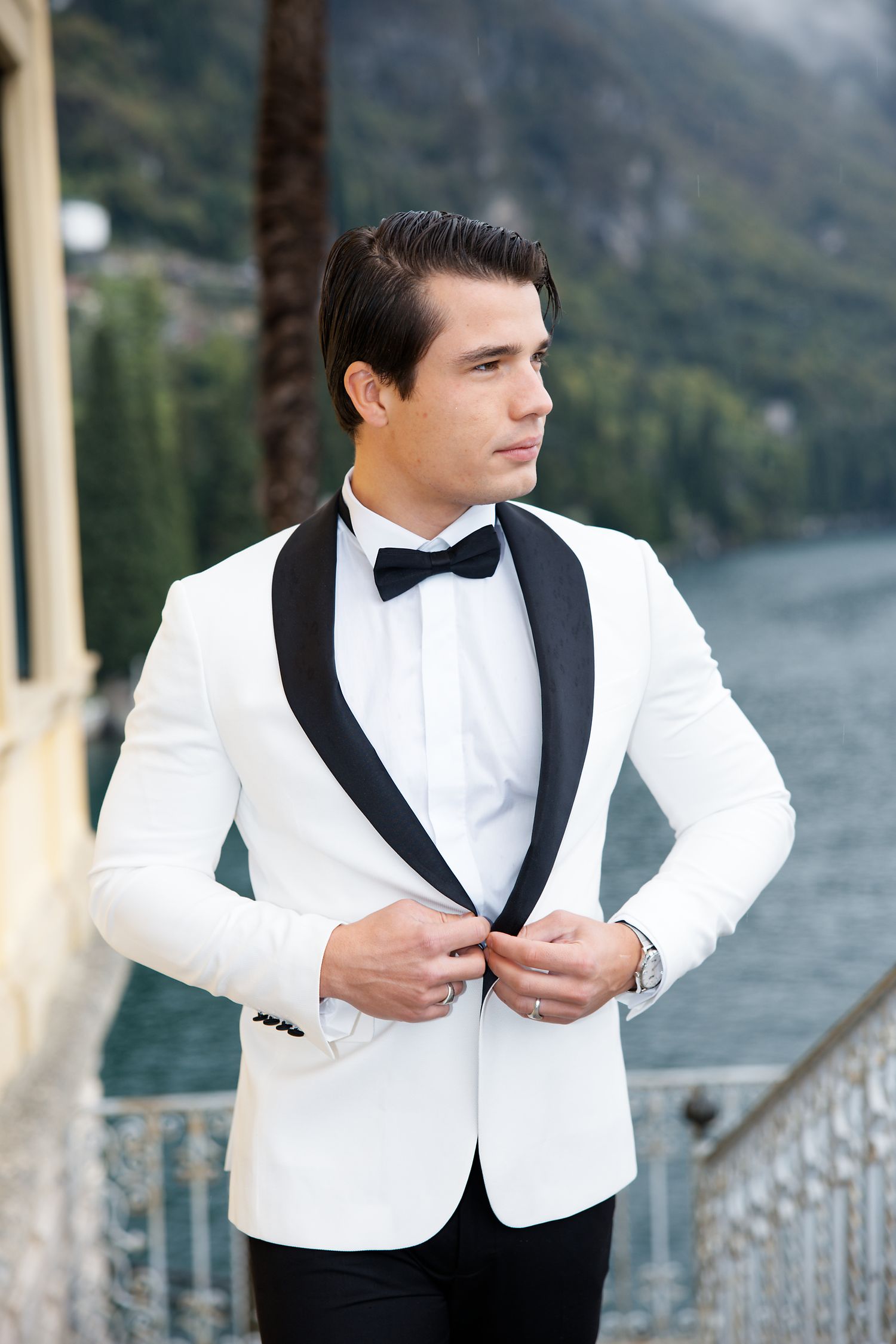Sophisticated groom adjusts white dinner jacket while standing on lakeside terrace.