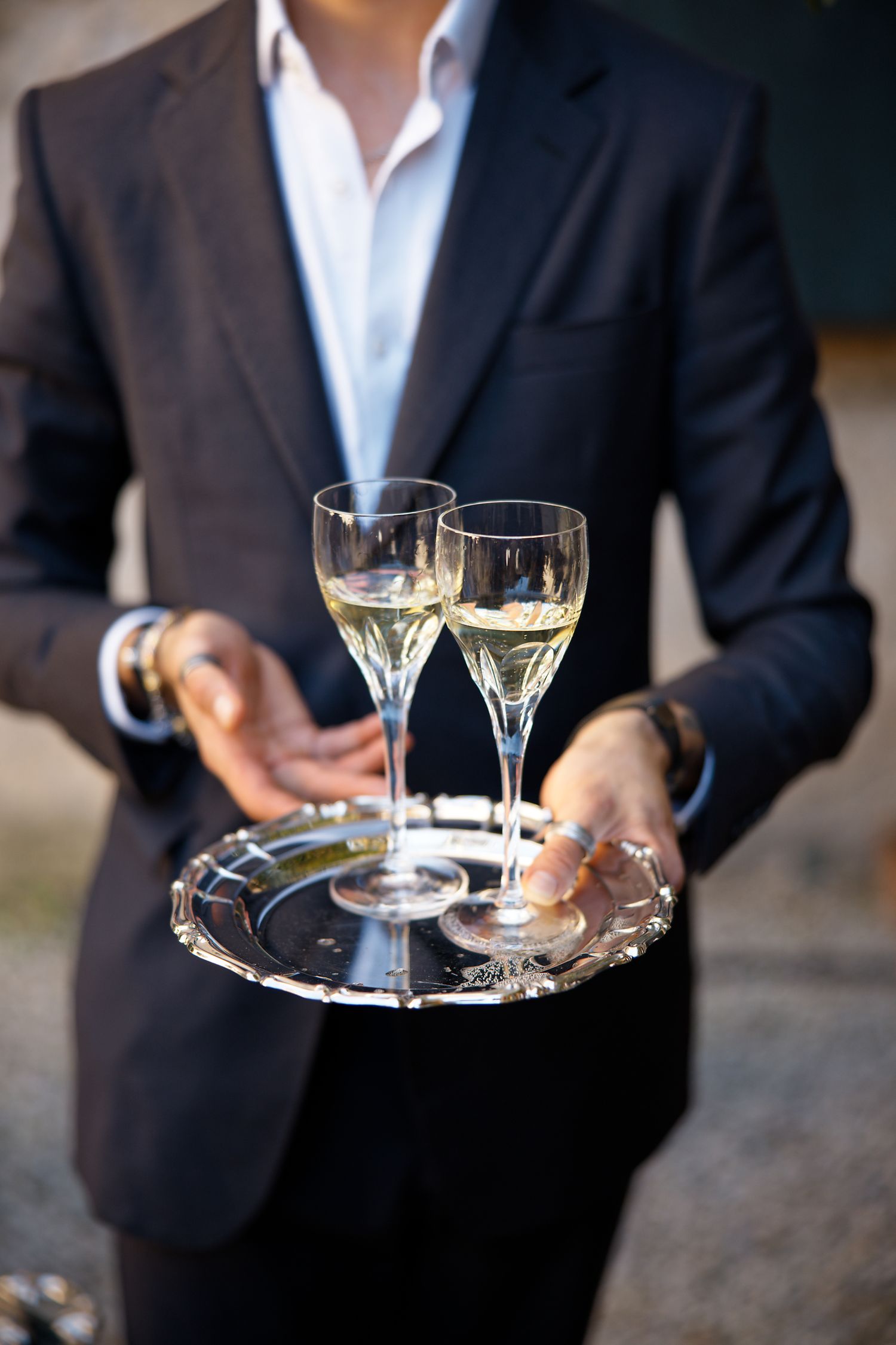 Server in black suit holding silver tray with two champagne glasses in elegant setting.