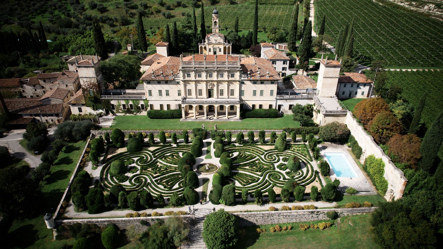 Aerial view of a luxurious Italian villa estate with manicured geometric gardens and swimming pool surrounded by vineyards.
