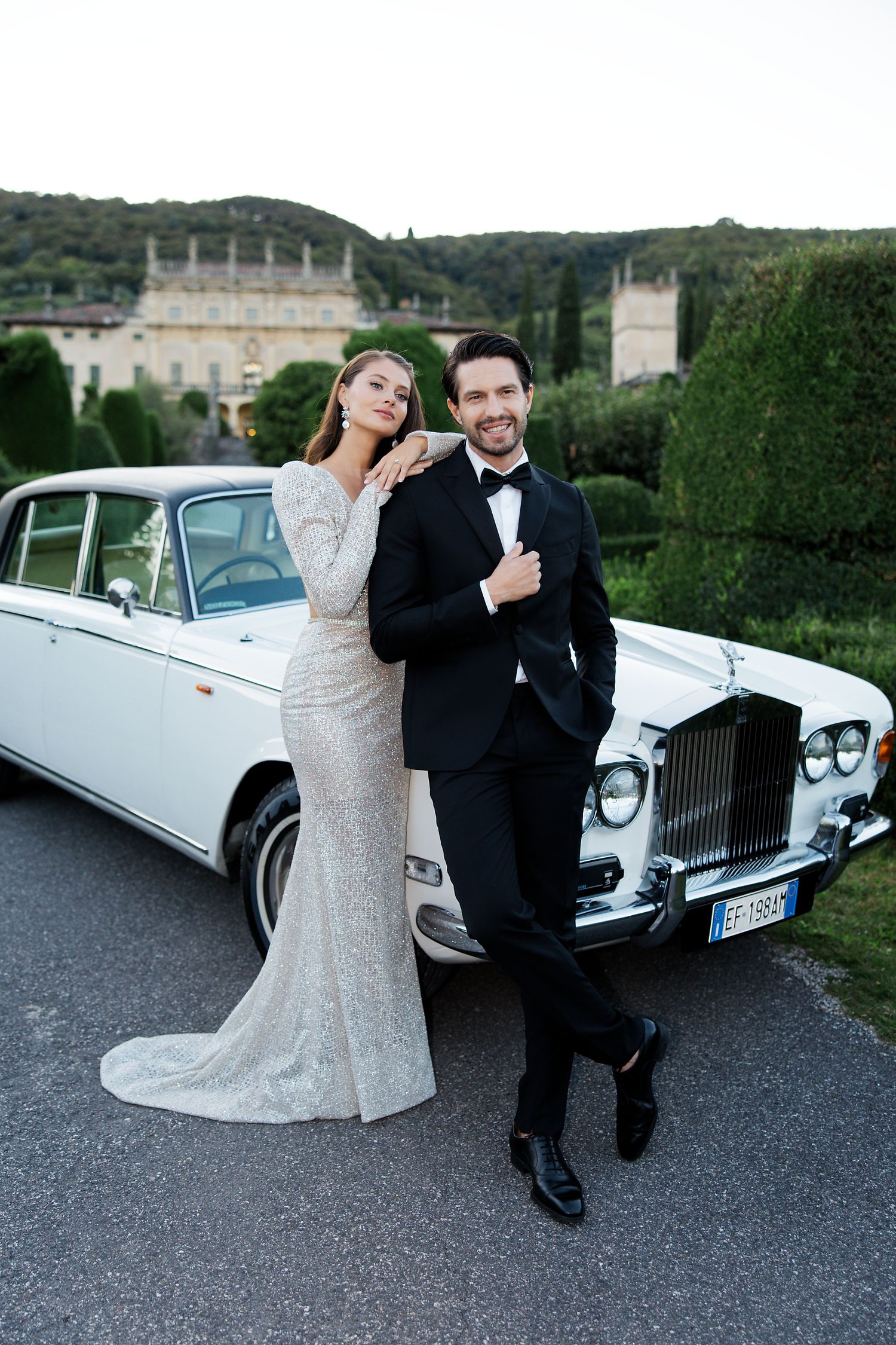 A classic white Rolls Royce parked outside an elegant mansion with a couple in formal attire.