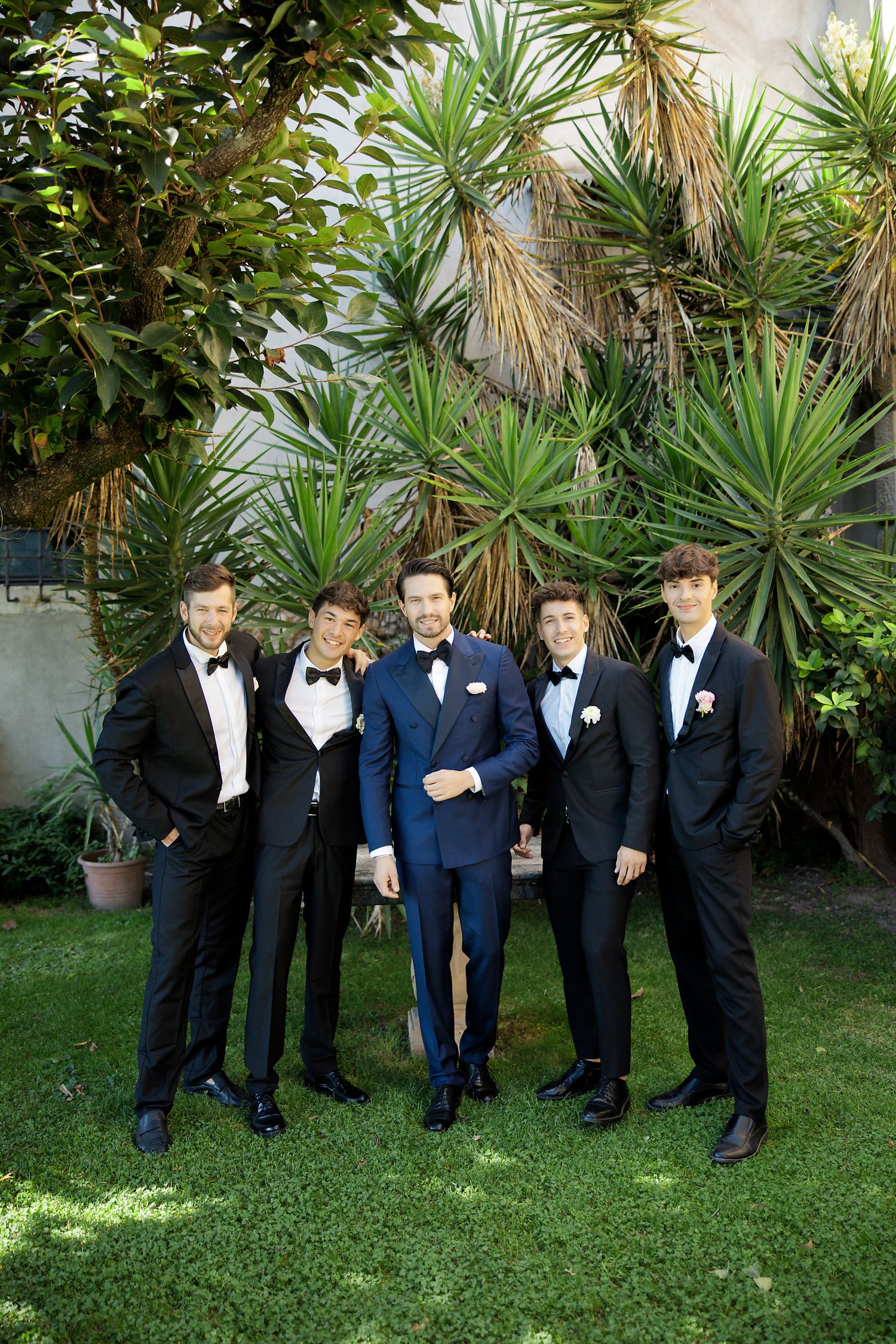 A formal wedding party wearing tuxedos poses together on grass in front of tropical plants and palm trees.