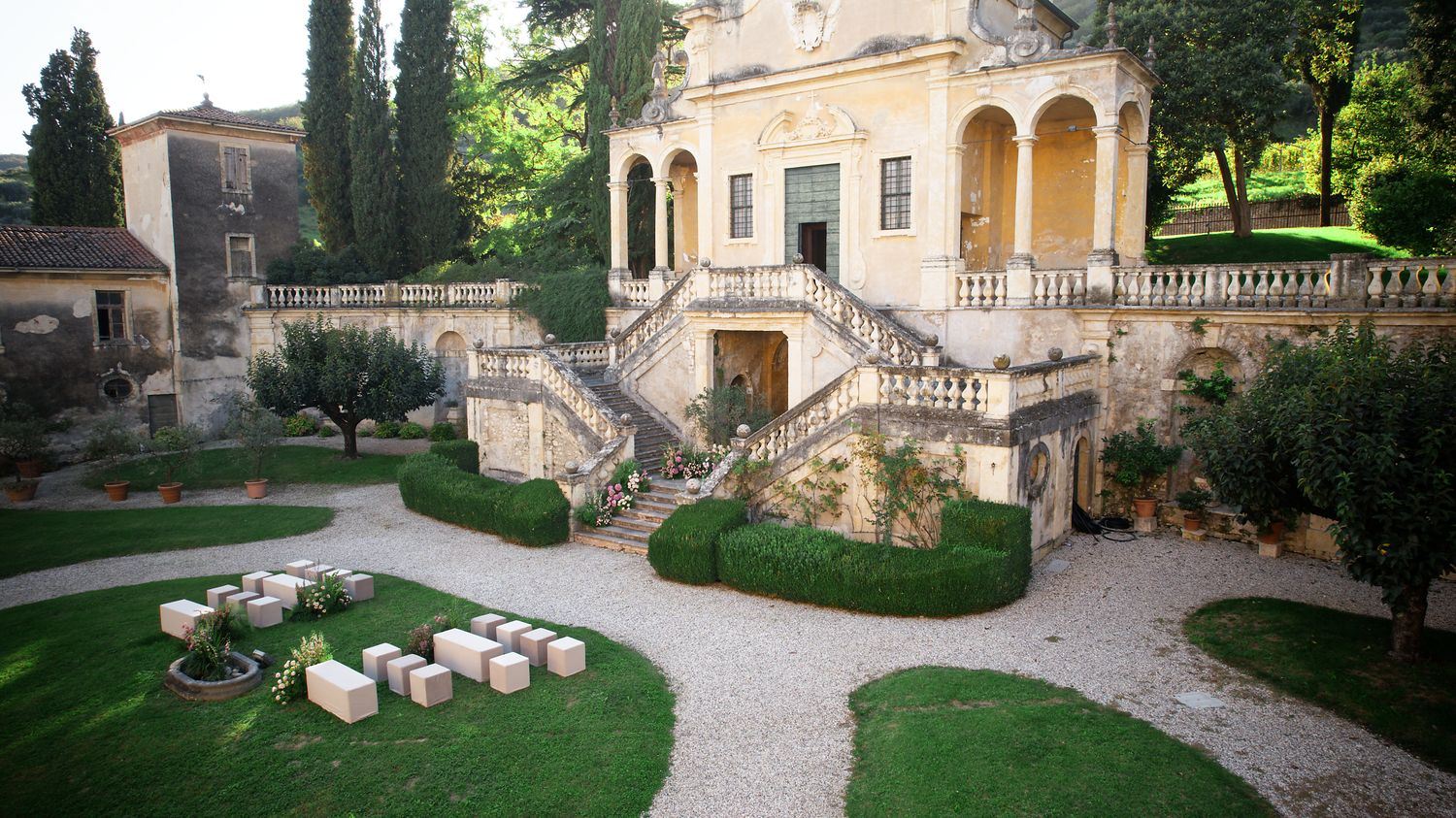 A Mediterranean villa courtyard with curved stone stairs, manicured lawns, and gravel pathways.