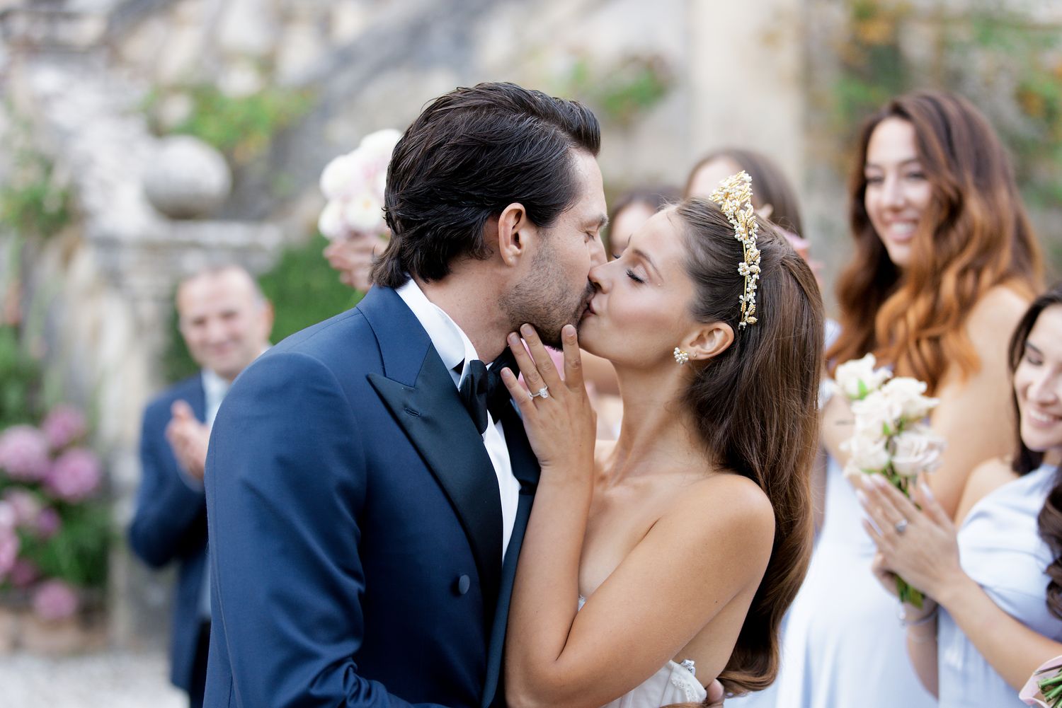 Newlyweds share a romantic kiss at an outdoor wedding ceremony while guests applaud in the background.