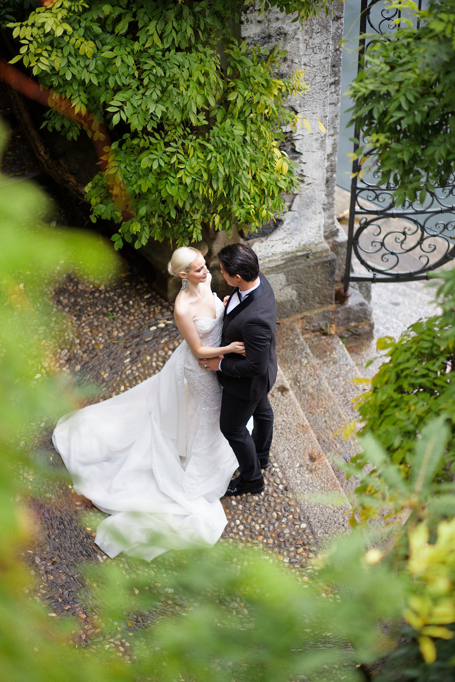 Wedding couple sharing intimate moment on stone terrace surrounded by lush greenery and decorative railings.