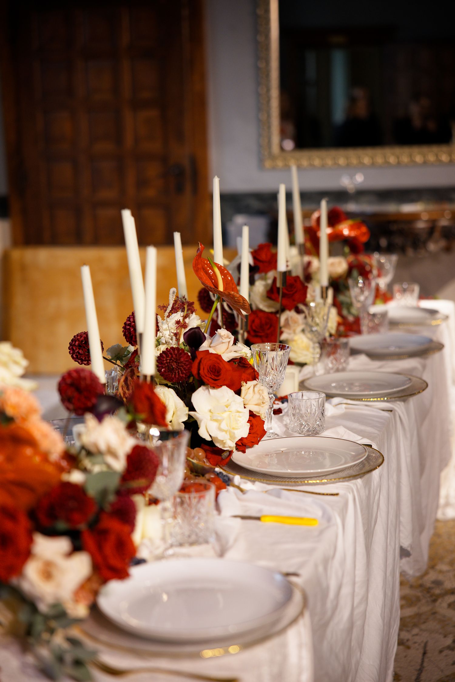 A formal dining table decorated with red and white flowers, candles and fine china place settings.