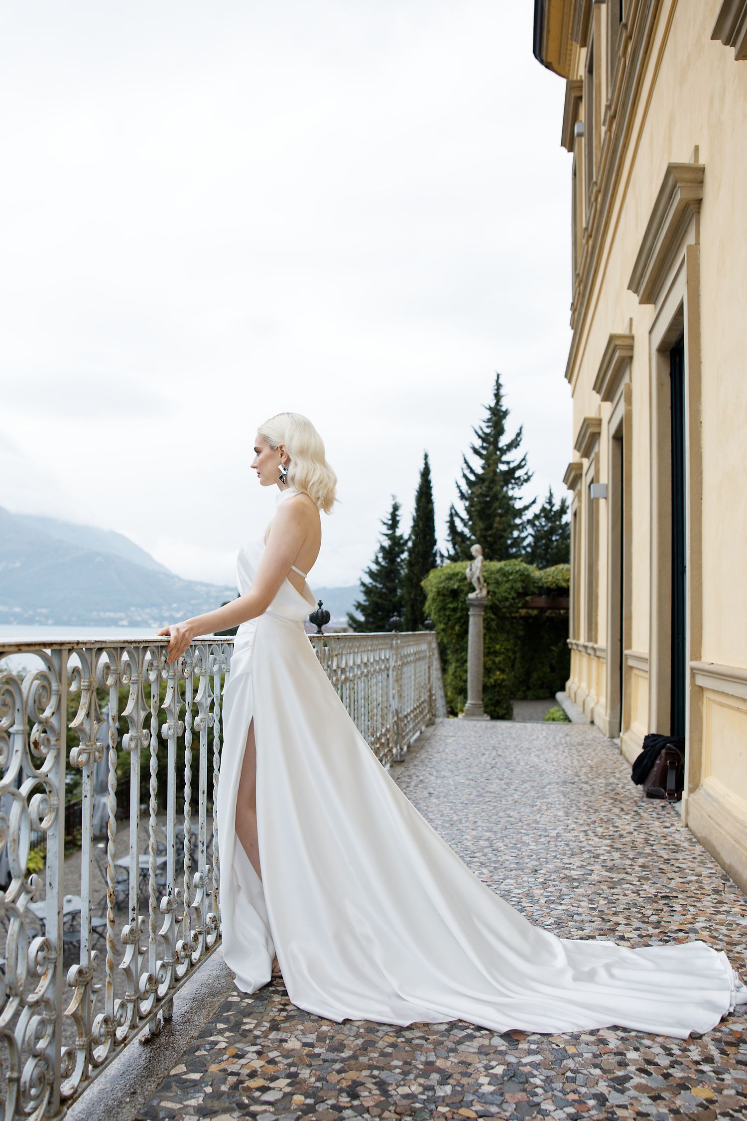 Bride in flowing white gown with train stands on ornate balcony overlooking mountains.