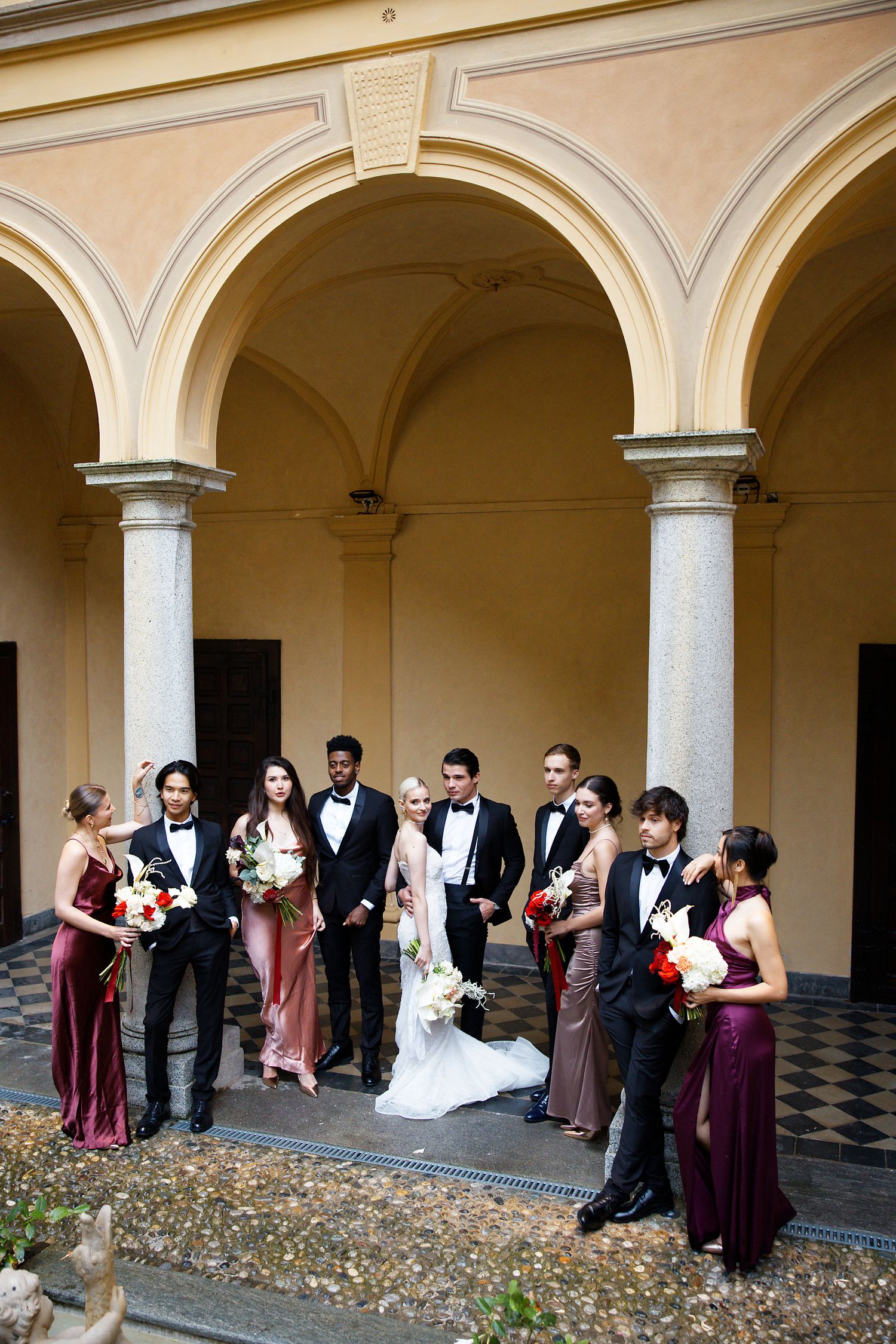 Wedding party in formal attire poses under arched colonnade during elegant celebration.