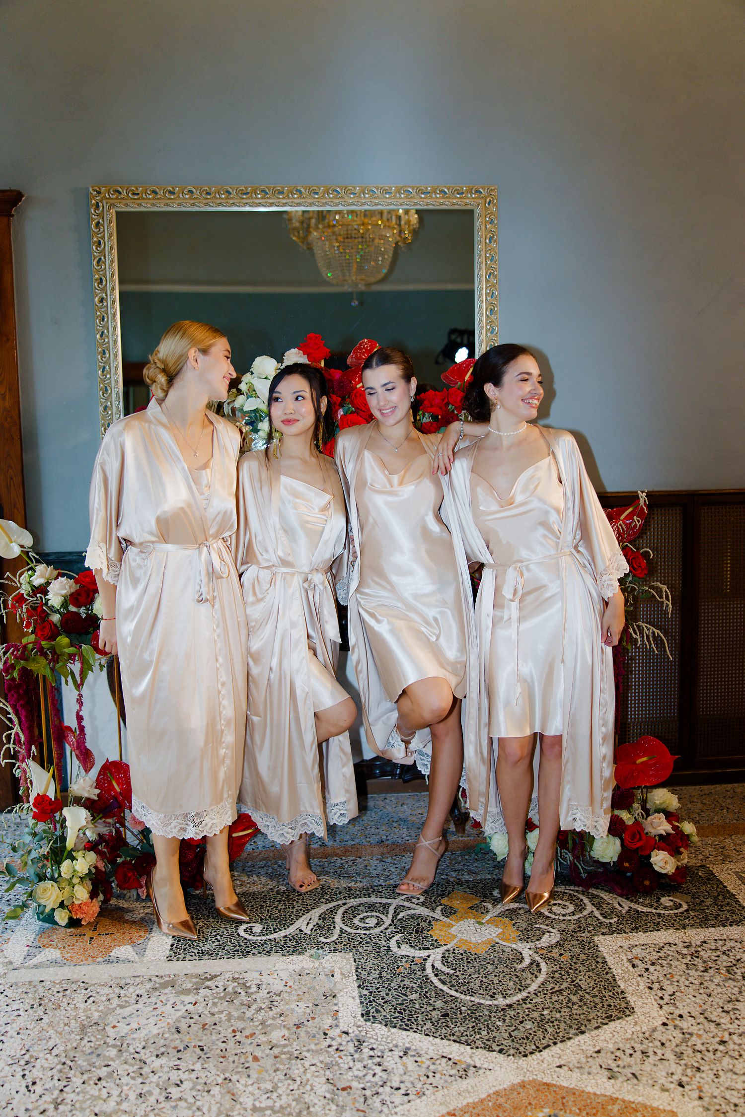 Four bridesmaids posing together in silk robes surrounded by red floral decorations during wedding preparations.