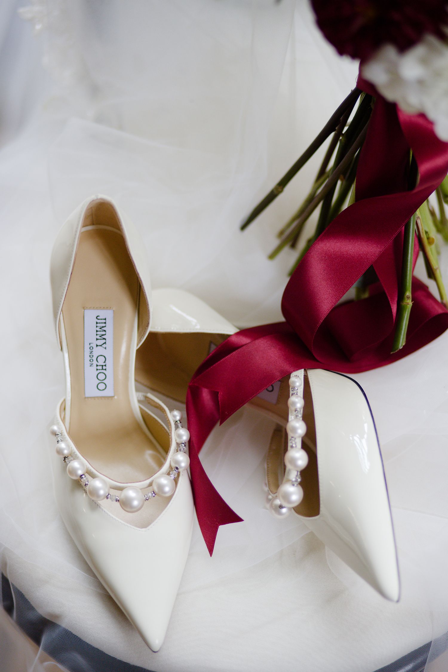 Elegant white Jimmy Choo bridal shoes with pearl detailing displayed next to deep red roses and ribbons.
