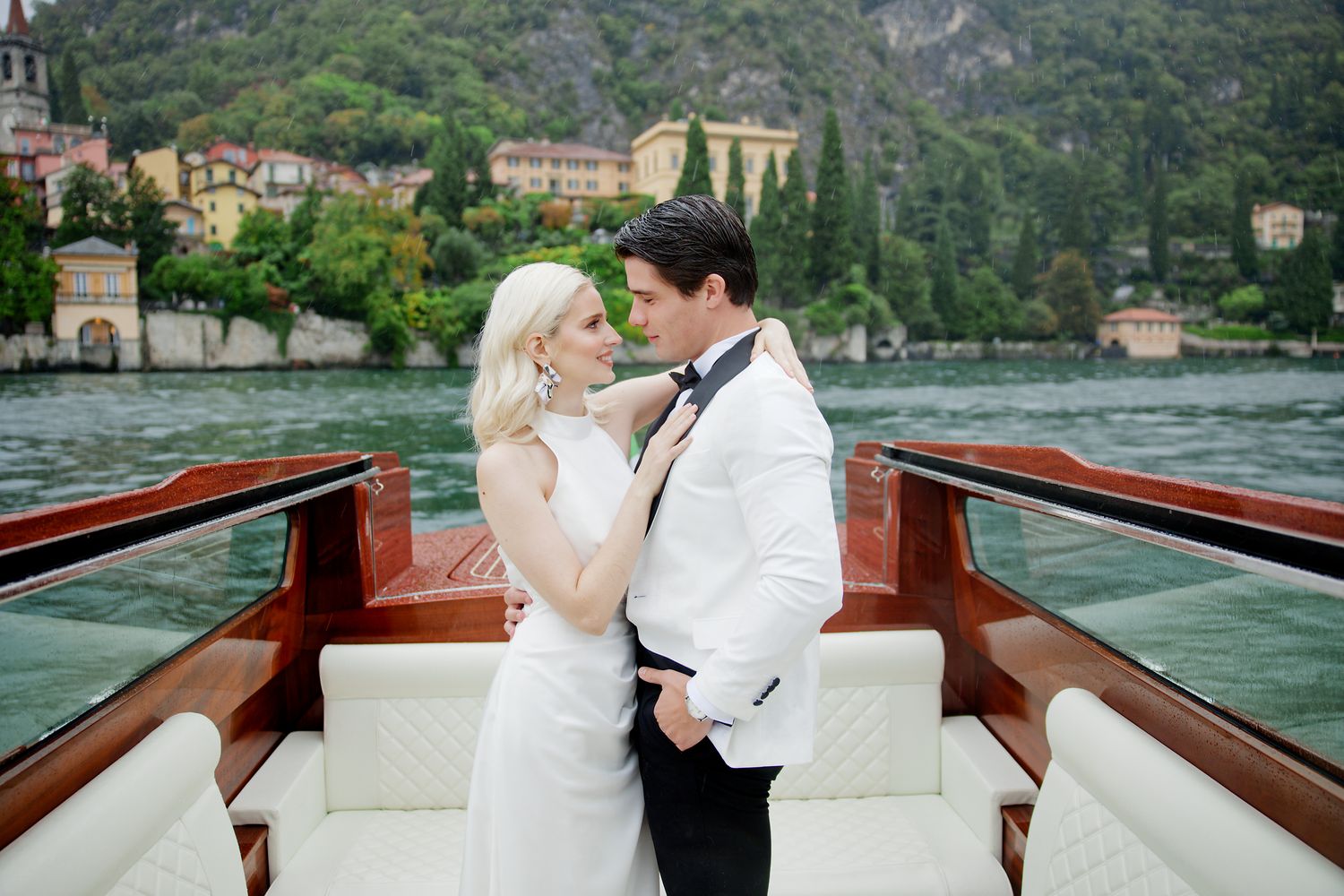 Romantic newlywed couple embracing on vintage wooden boat with Italian coastal town in background.