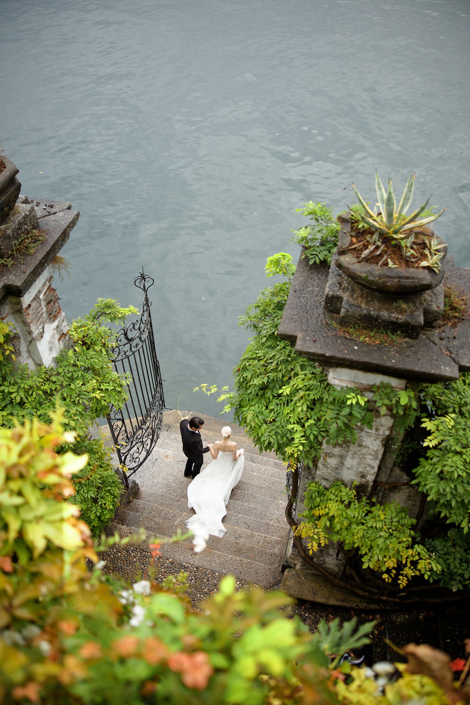 Aerial view of couple at lakeside villa steps surrounded by lush greenery.
