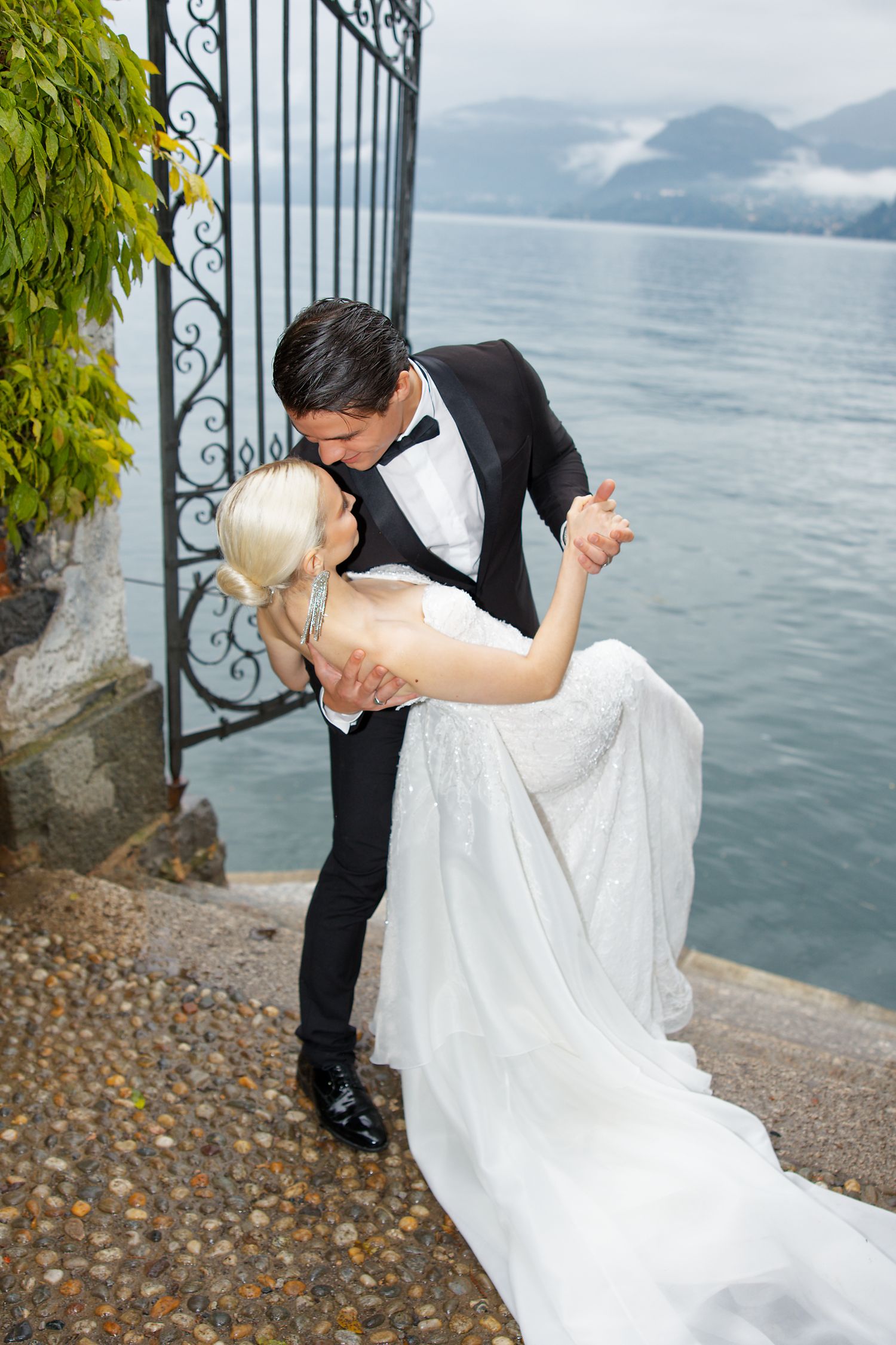 A romantic dip pose on a lakeside terrace with wrought iron gates and mountain views.