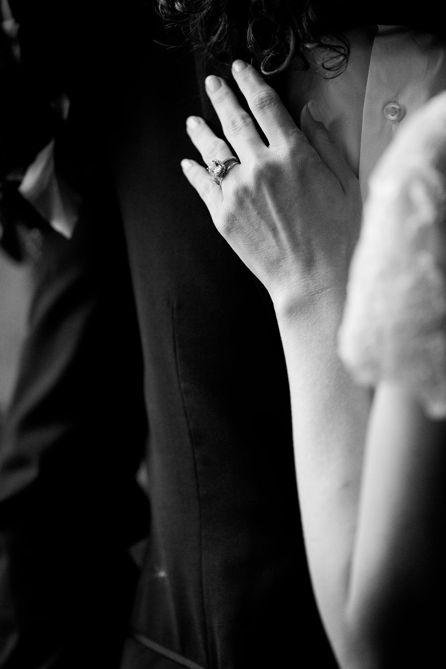 Close-up black and white photograph showing a diamond ring on a hand against a dark background.