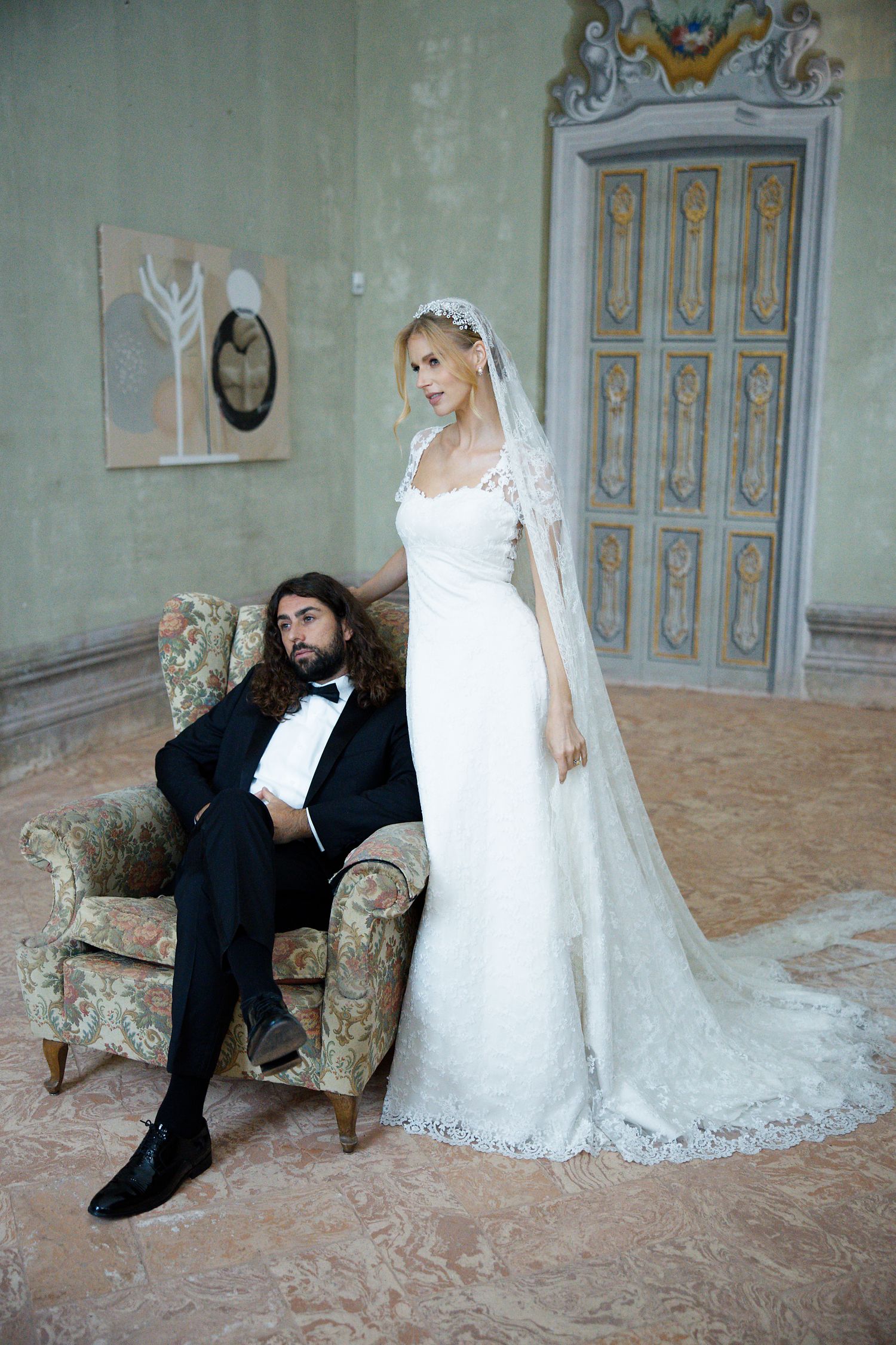 A wedding portrait featuring a long white lace gown with veil and ornate vintage armchair in an elegant setting.